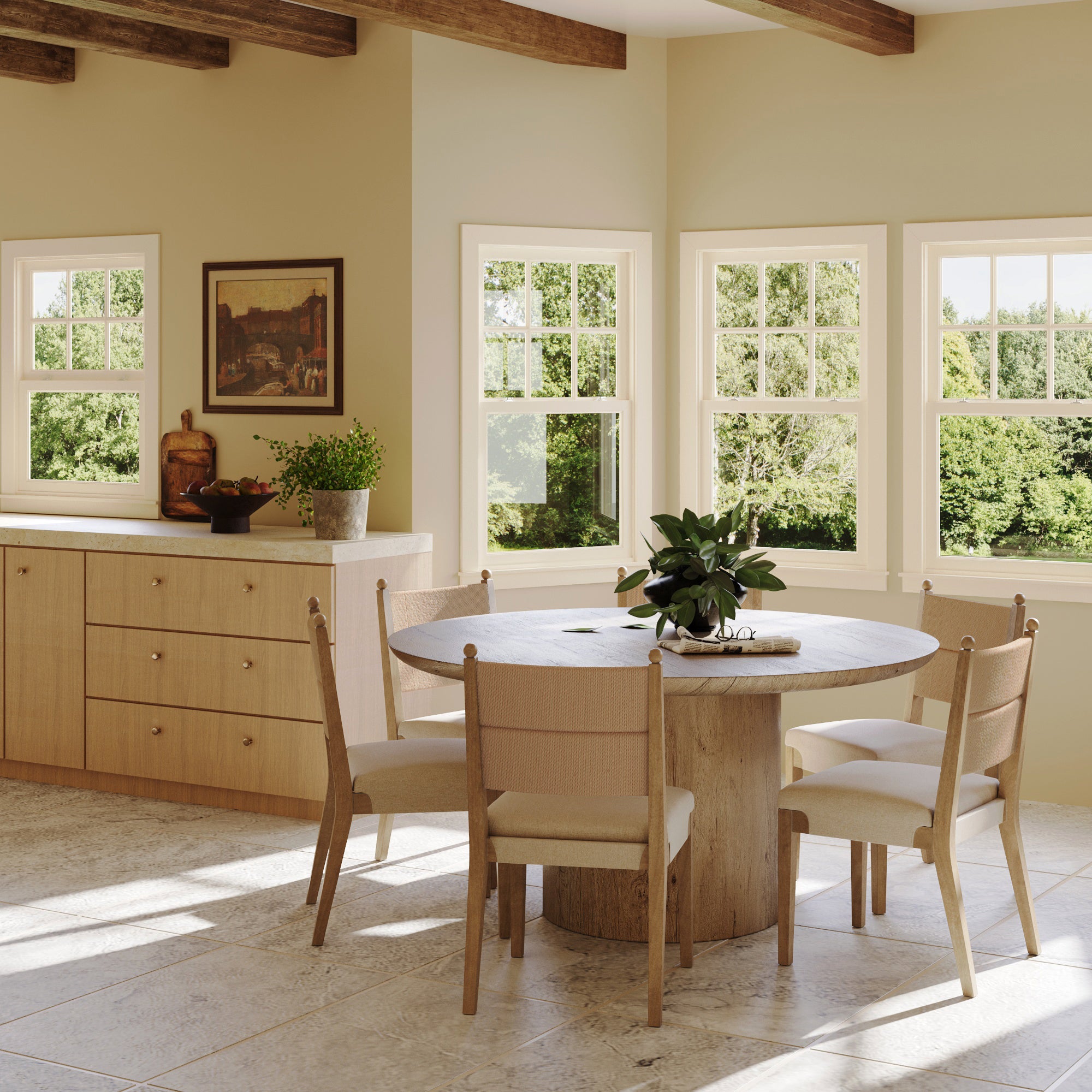 A bright, modern dining area features a round wooden table, four Nathan James Boho Natural Flax & Seagrass Dining Chairs, beige walls, large sunlit windows, green plants, and wooden beams for a warm and inviting atmosphere.
