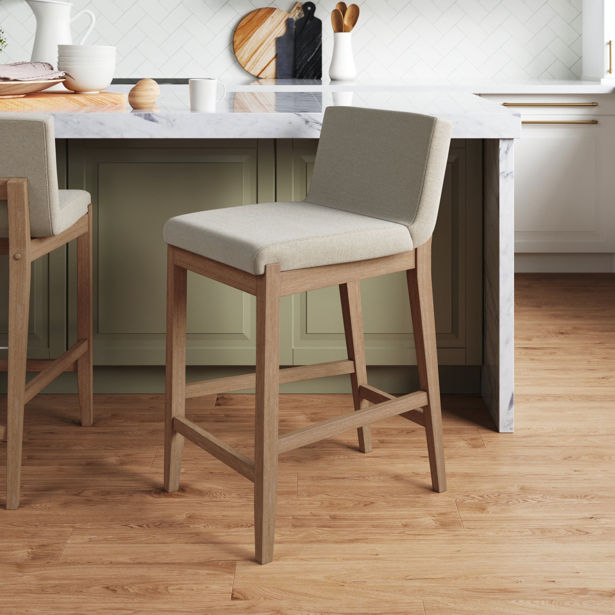 A light wood and beige upholstered bar stool stands in front of a marble kitchen island with sage green cabinets. The background features a herringbone tile backsplash, wooden cutting boards, and various kitchen items.