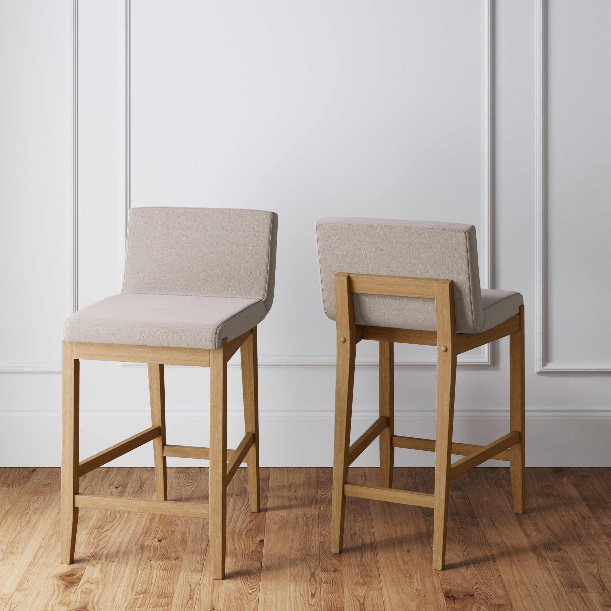 The Nathan James Wood Counter Height Bar Stools Light Brown (Set of 2) feature light beige upholstered seats and backs with wooden legs. Placed on a wooden floor by a white wall, one stool faces forward; the other shows the back.