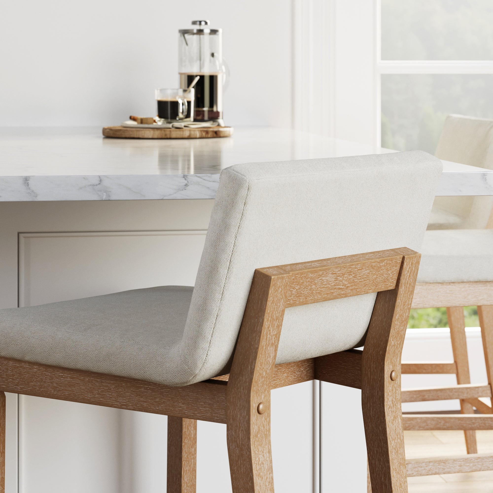A light wood and beige upholstered chair sits by a marble countertop. On the counter are a French press, a glass of coffee, and a wooden tray. A large window provides natural light in the background.