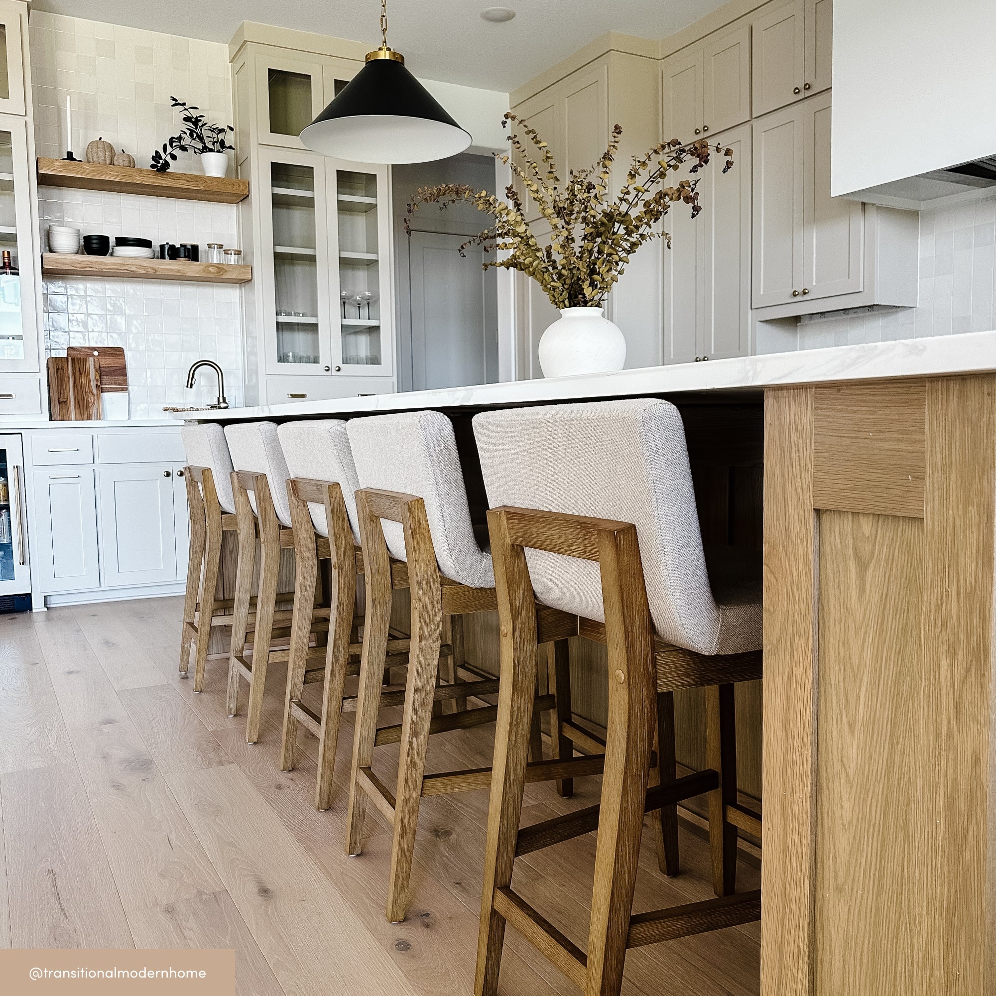 A modern kitchen with light wood floors, a long island with wood and upholstered barstools, white cabinets, black pendant light, and a white vase with foliage on the countertop.