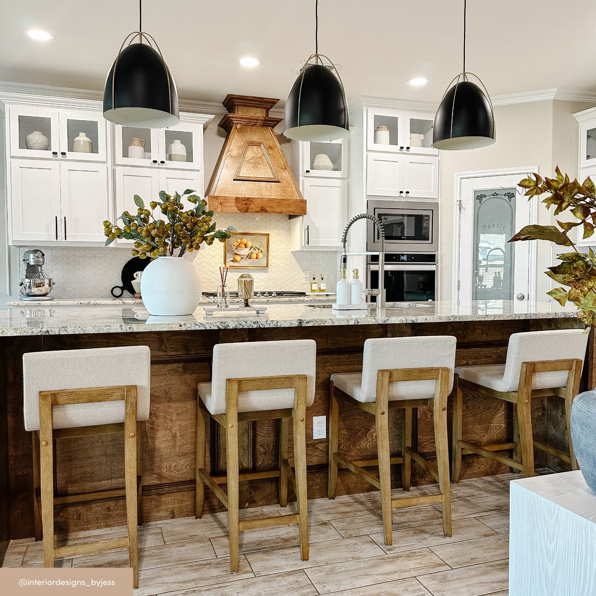 A modern kitchen with a large island, four wooden barstools with cream cushions, white cabinetry, stainless steel appliances, and three black pendant lights hanging above the island. A large vase with greenery sits on the counter.