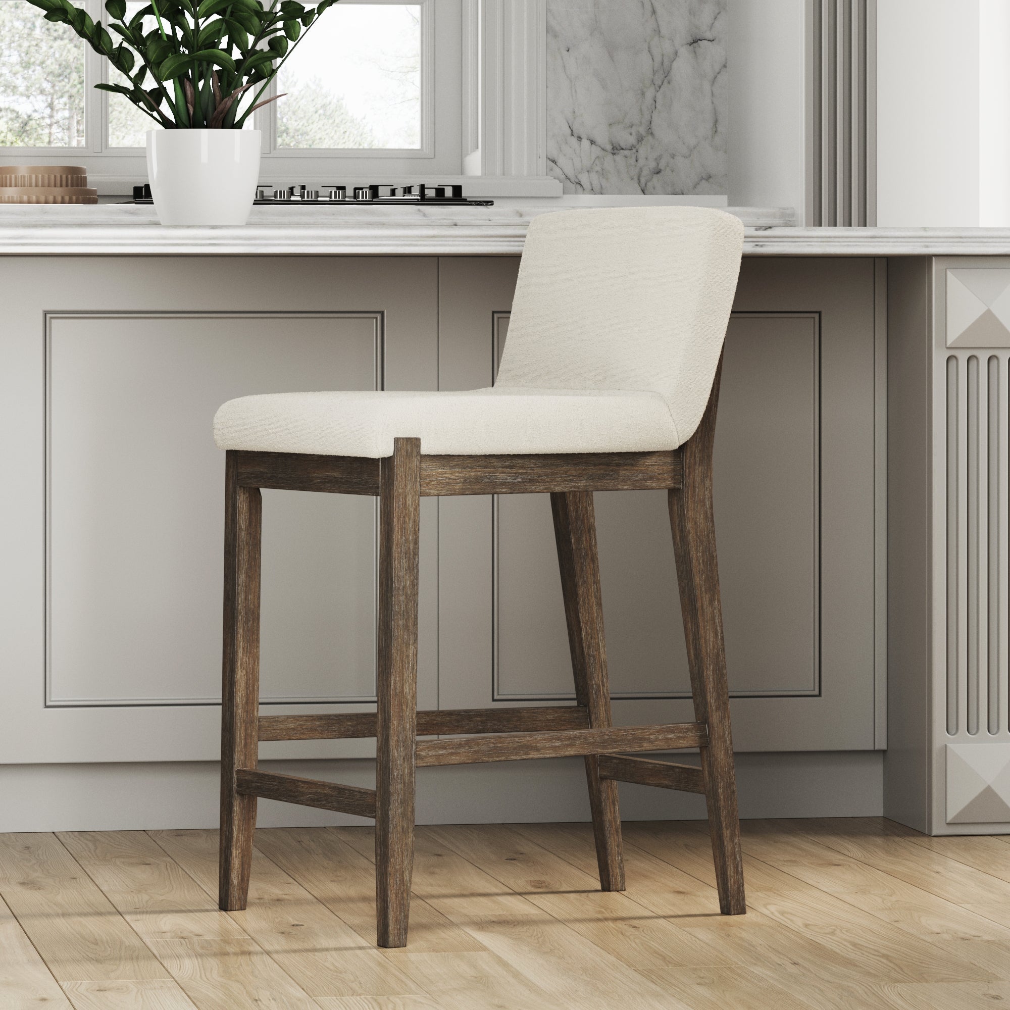 A Nathan James Wood Boucle Counter Height Bar Stool in dark brown, featuring a cushioned beige seat and backrest, stands on a light wood floor in a modern kitchen with gray cabinets, white countertop, and a potted plant by the window.