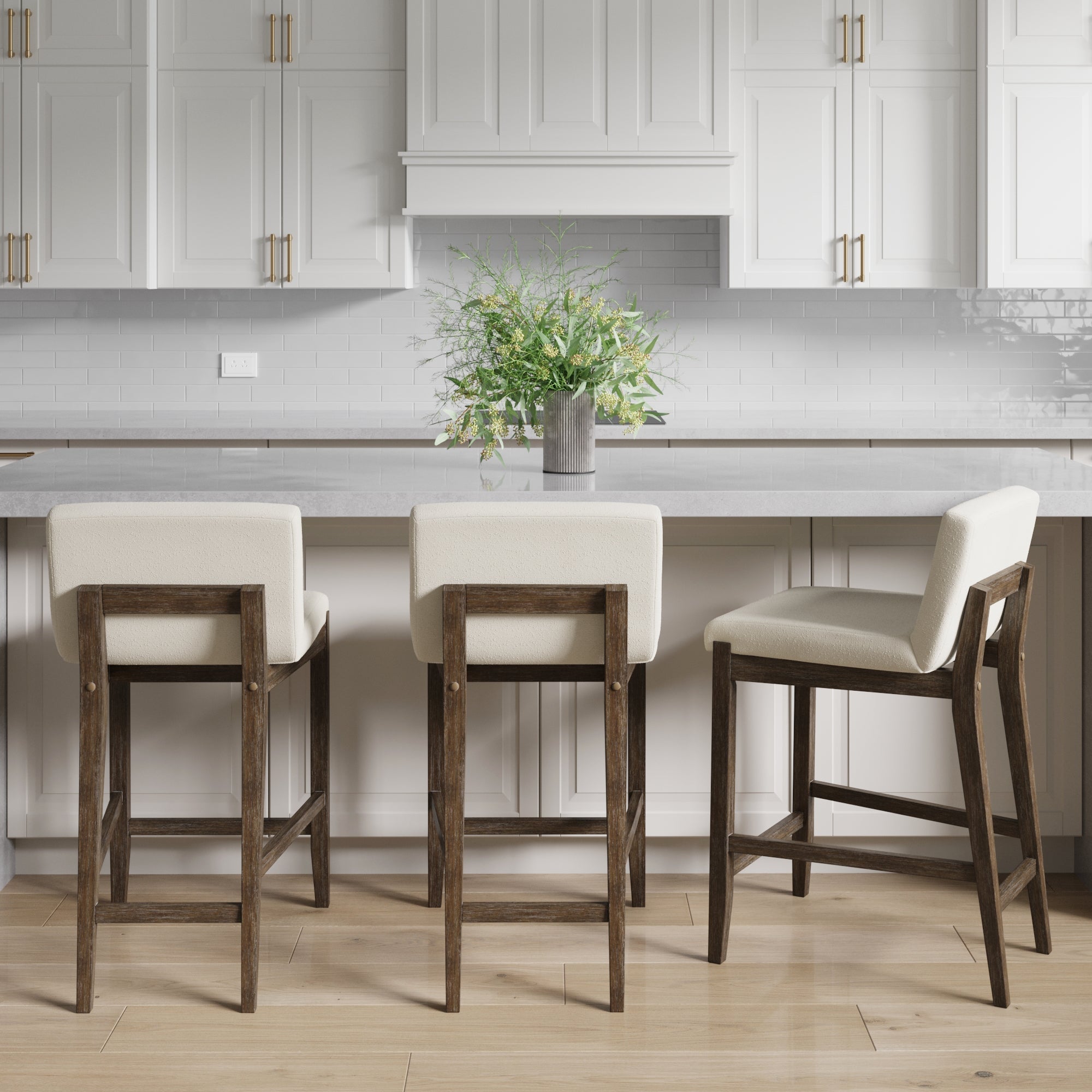 Three Nathan James Wood Boucle Counter Height Bar Stools in dark brown with white cushioned seats are arranged at a white kitchen island, with white cabinets, a tiled backsplash, and a potted plant on the counter in the background.