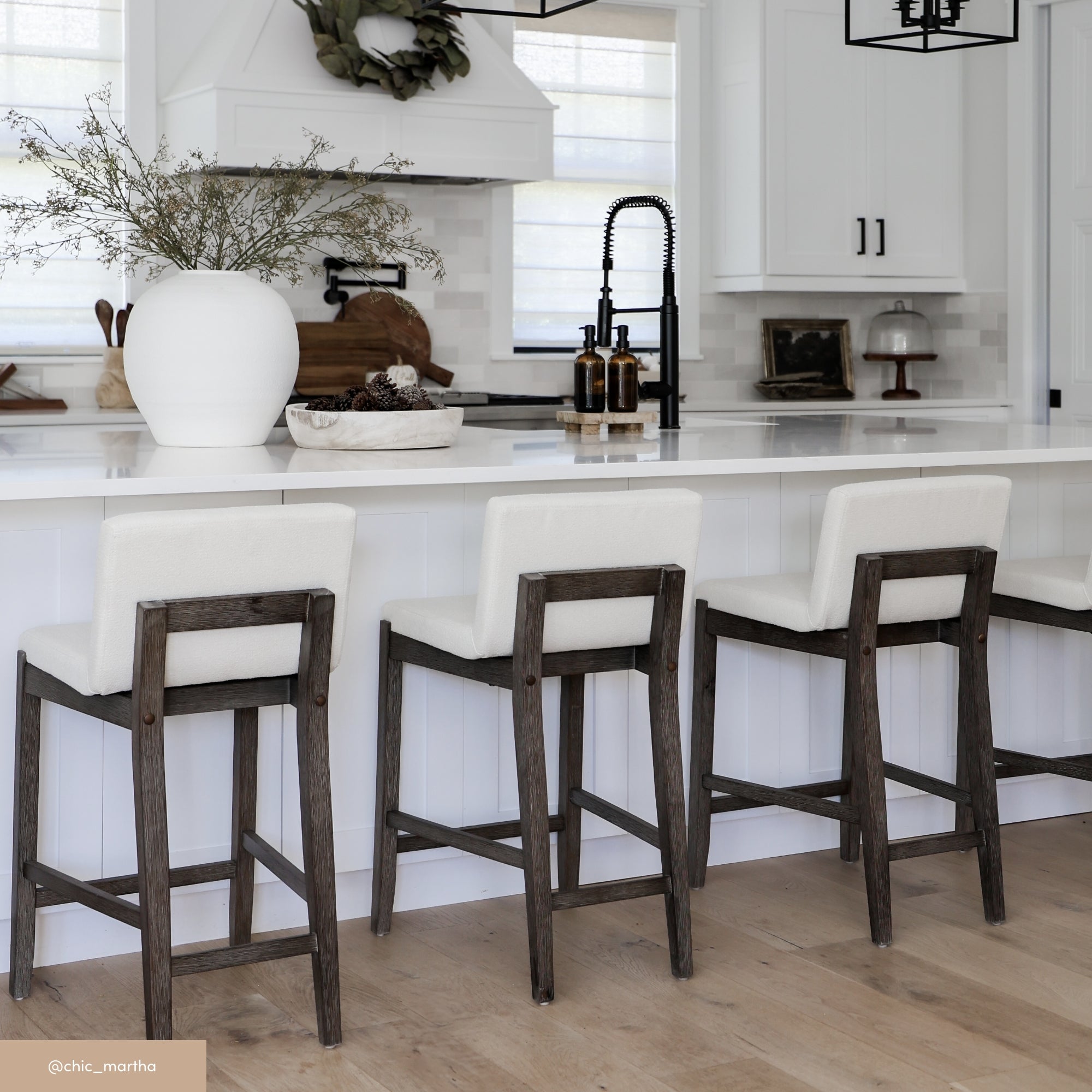 Modern kitchen with a white island, three cushioned bar stools with dark wooden frames, minimalist decor, a large white vase with greenery, and light wood flooring. White cabinets and black fixtures are visible in the background.