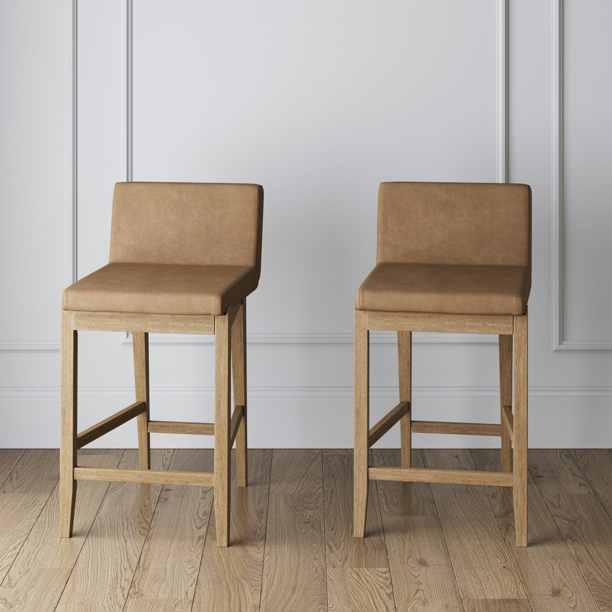 Two light brown barstools with cushioned seats and backs, featuring wooden legs, are placed side by side on a wooden floor against a white paneled wall.