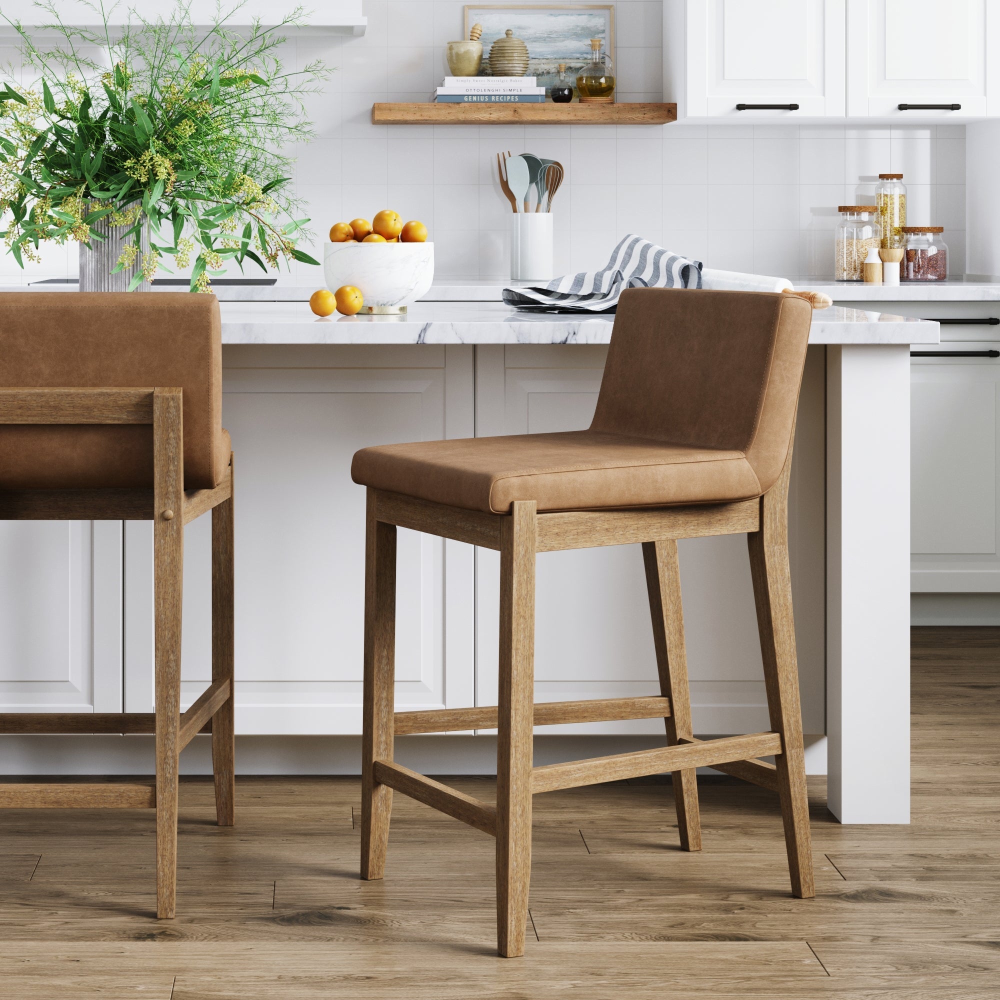 A modern kitchen features two Nathan James Wood Faux Leather Counter Height Bar Stools in light brown at a marble island, surrounded by white cabinets, a bowl of oranges, greenery in a vase, and decorative items on shelves.