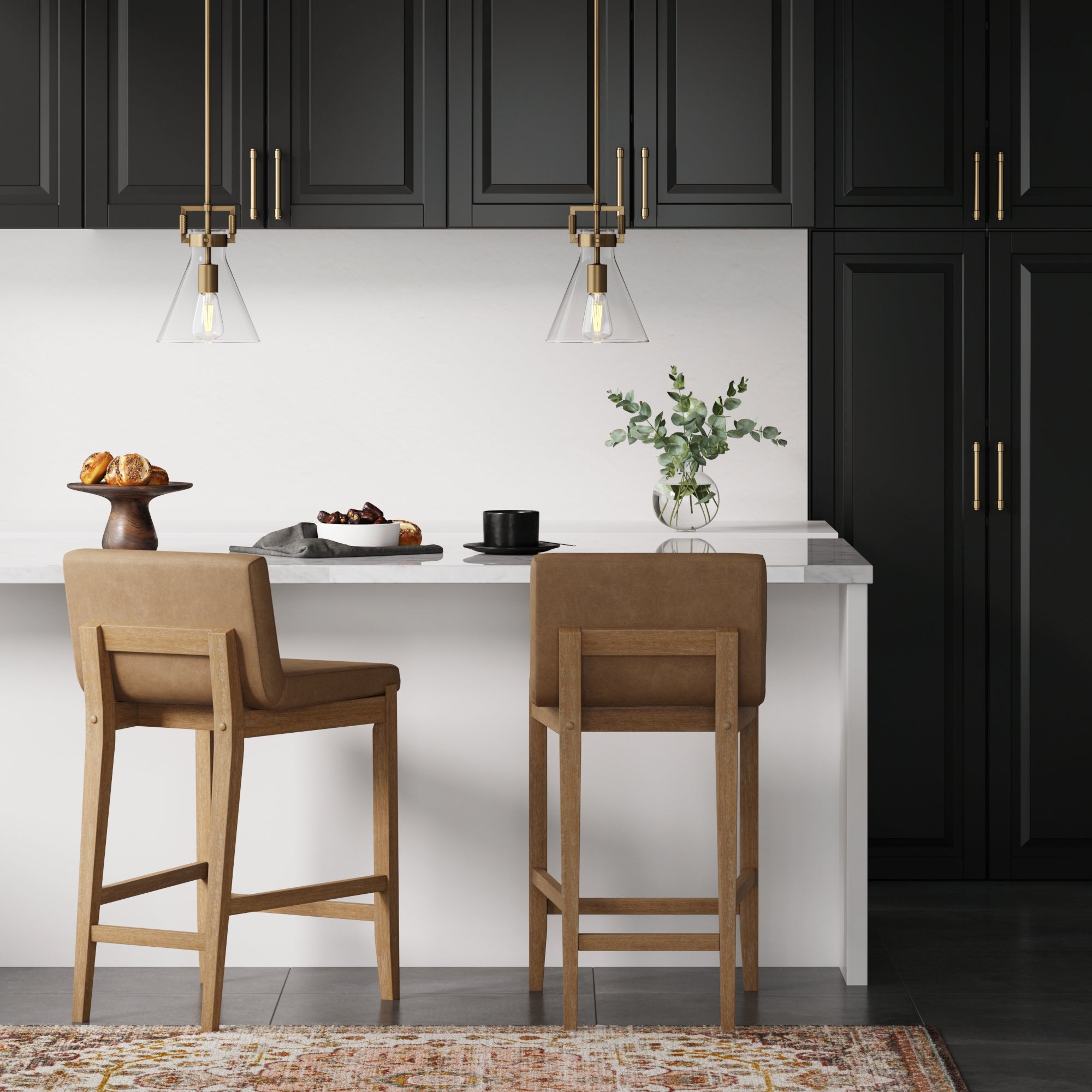 A modern kitchen with black cabinets, gold hardware, a white countertop, two brown bar stools, hanging pendant lights, pastries on a cake stand, a coffee cup, a plant in a vase, and a patterned rug.