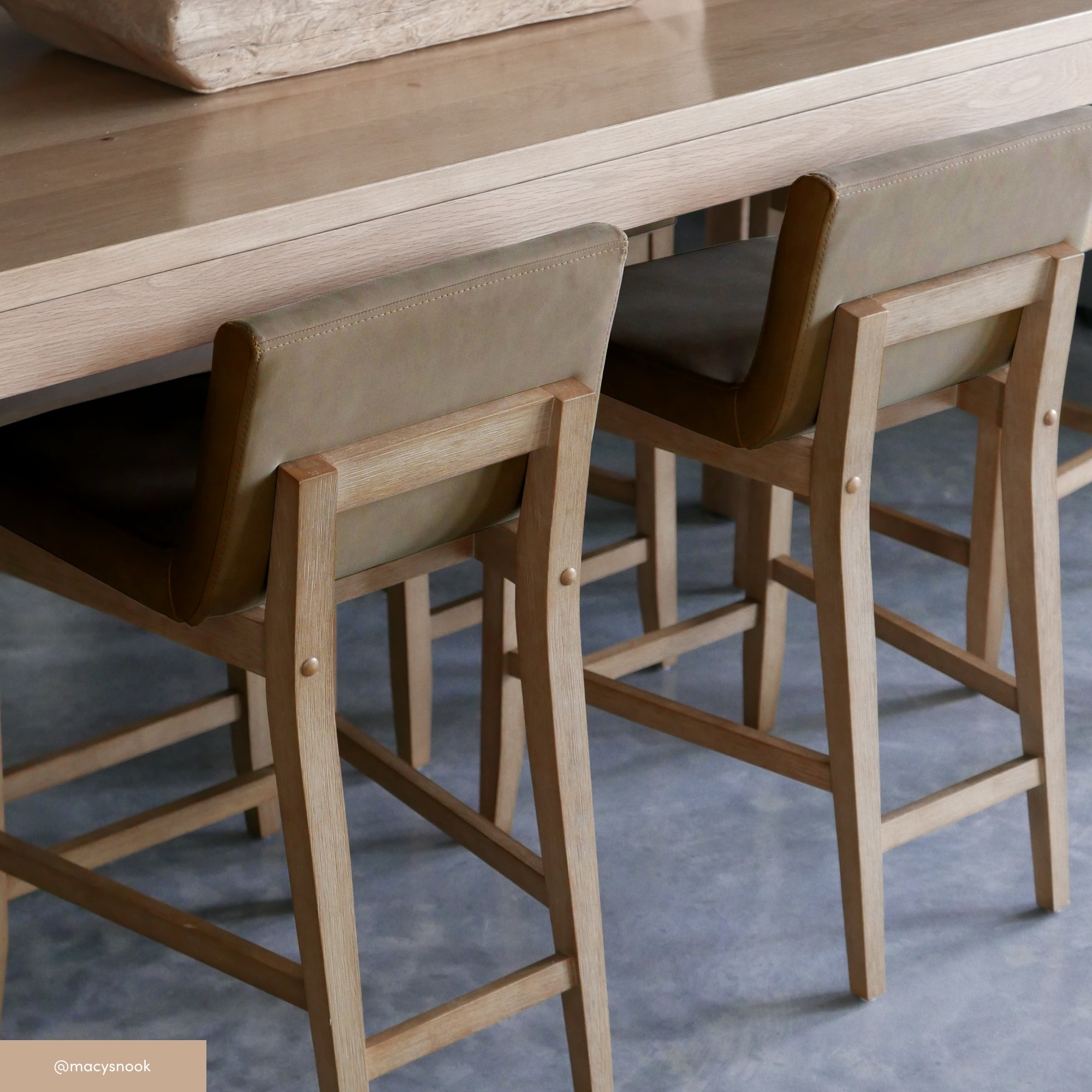Two light wooden barstools with olive green cushioned backs are placed at a matching wooden counter on a gray concrete floor. The overall style is modern and minimalist.