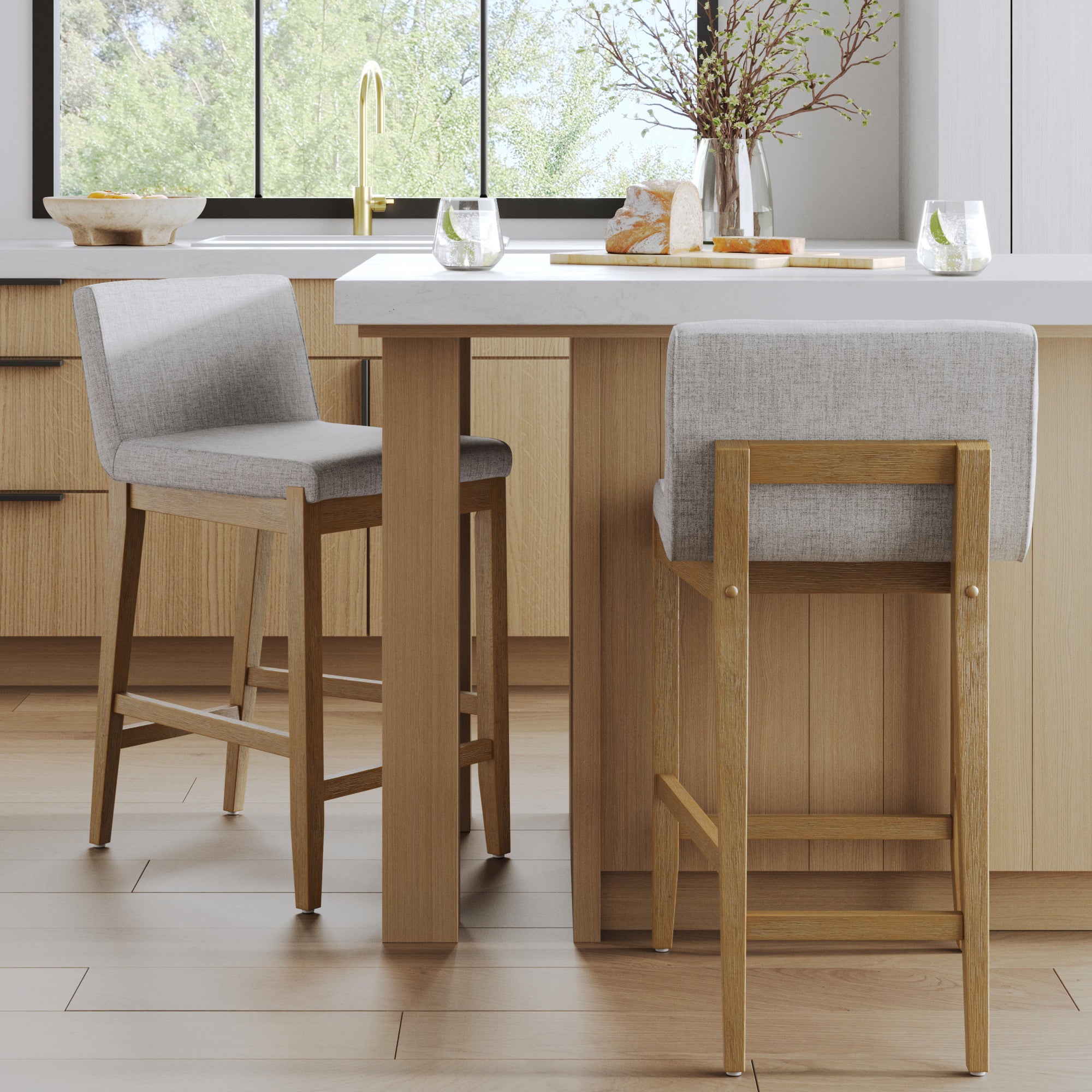 A modern kitchen with light wood cabinets features two Nathan James Upholstered Counter Height Bar Stools with gray seats at a white island, accented by glasses and a vase with branches for a stylish touch.