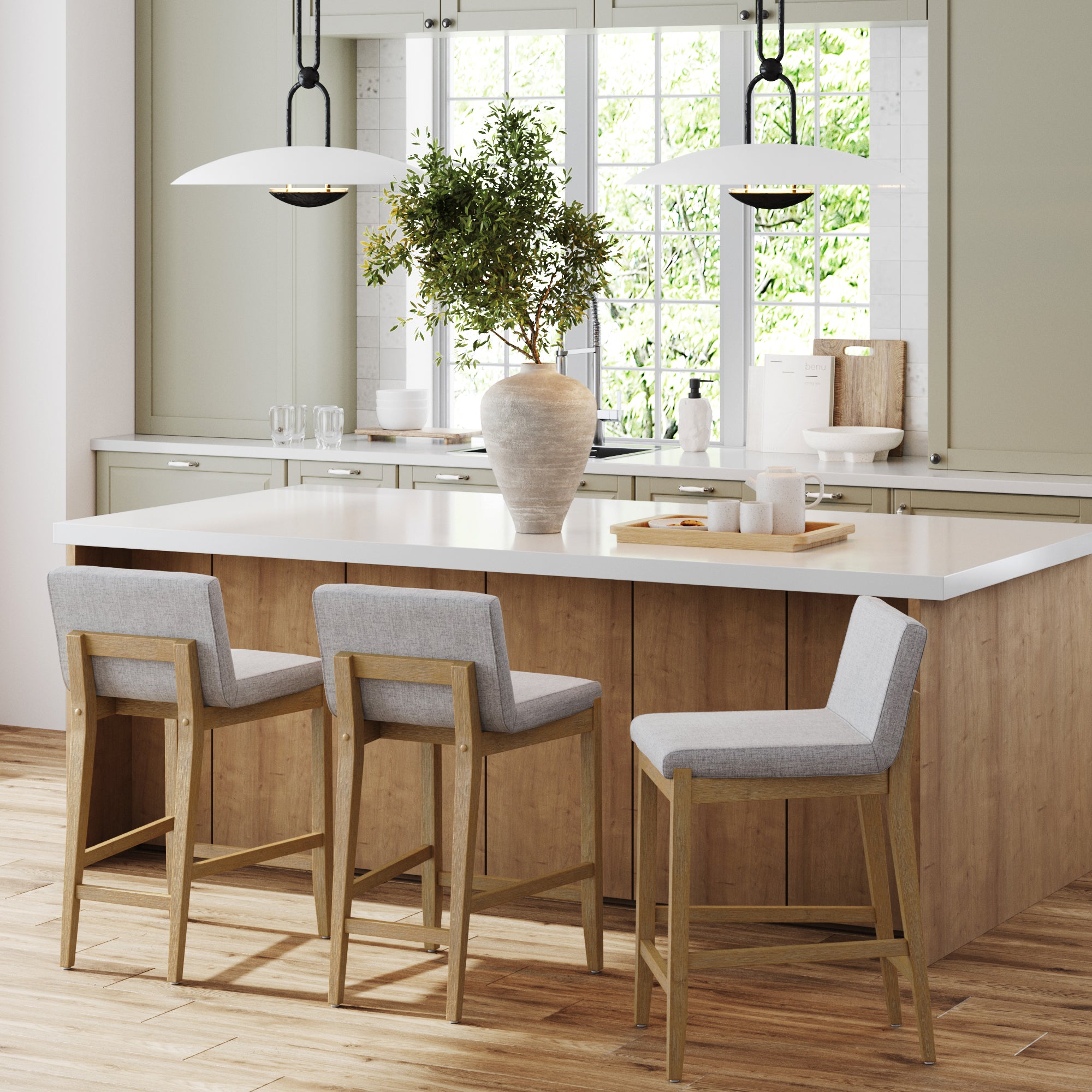 A modern kitchen features a large island with a white countertop, potted plant, pendant lights, window, and three Nathan James Upholstered Counter Height Bar Stools w/ Back in gray.