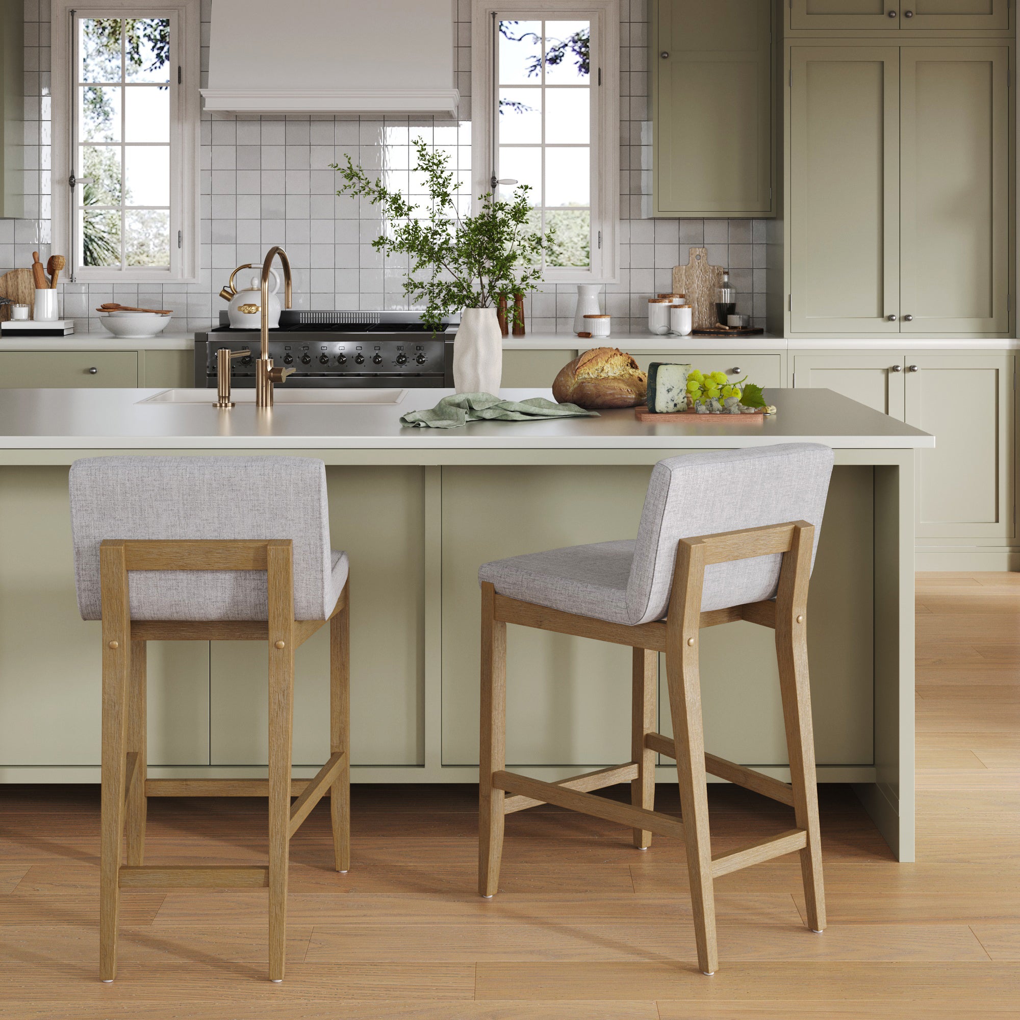 A modern kitchen features light green cabinets, a central island with two Nathan James Modern Upholstered Counter Height Bar Stools in gray, a gold faucet sink, countertops with bread, fruit, and a vase of greenery, and sunlight streaming through two windows.