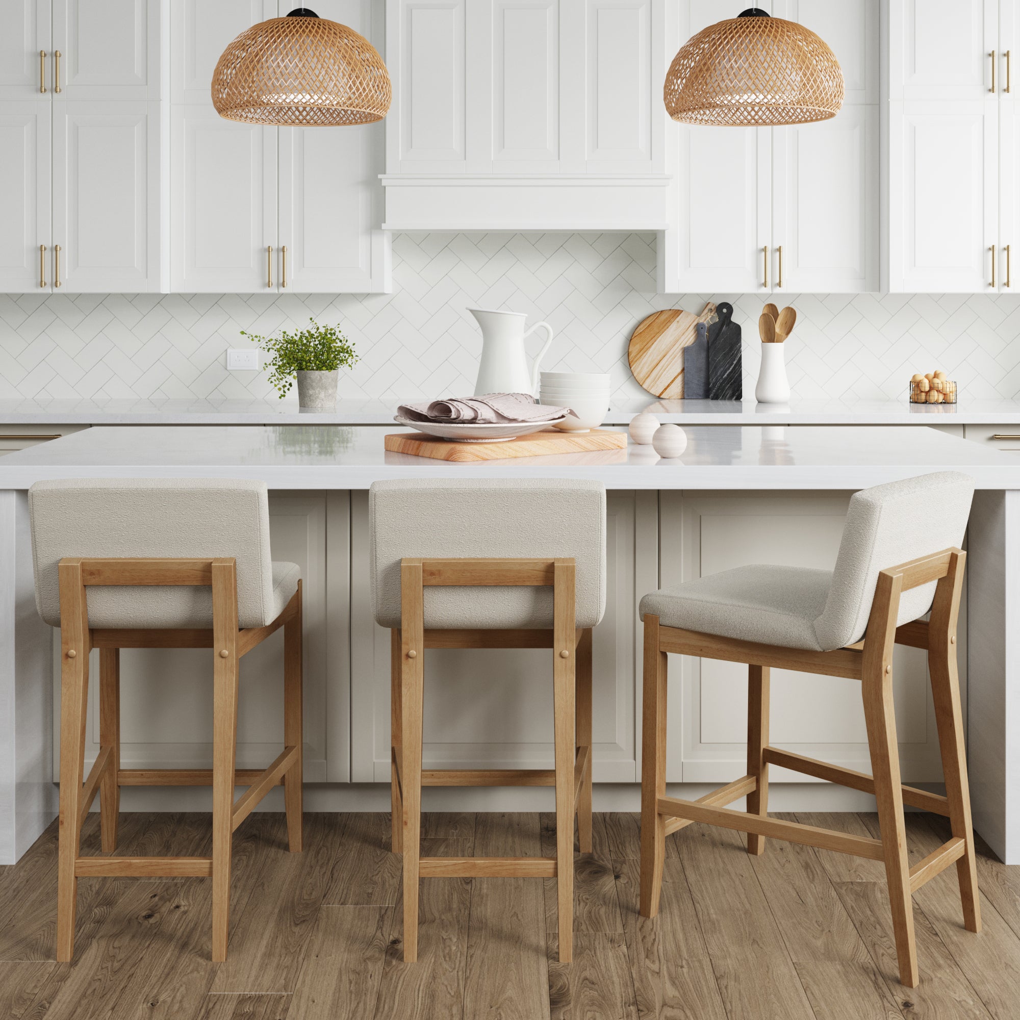 A bright modern kitchen features white cabinets, a white backsplash, and a central island with three Nathan James Boucle Counter Height Bar Stools in Warm Pine. Two wicker pendant lights hang above the island, with cutting boards and plants on the countertop.