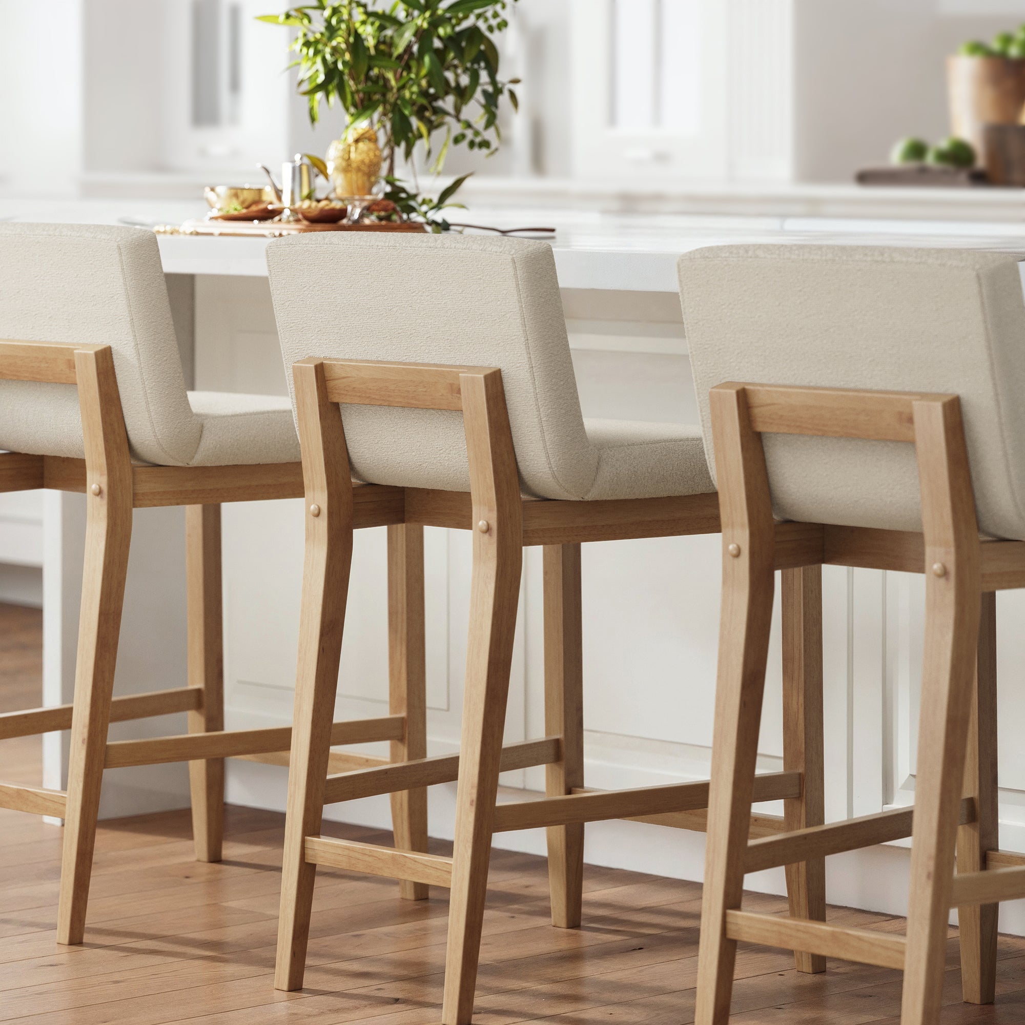 Three wooden bar stools with light beige upholstered seats are lined up at a white kitchen island. The kitchen features natural light, wooden flooring, and a small plant on the countertop.
