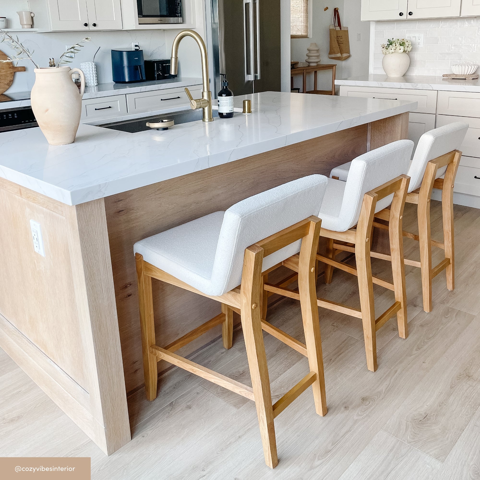 A modern kitchen with a large island featuring a white countertop, light wood base, and three wooden barstools with white seats. The kitchen has white cabinets and neutral decor, creating a bright, airy atmosphere.
