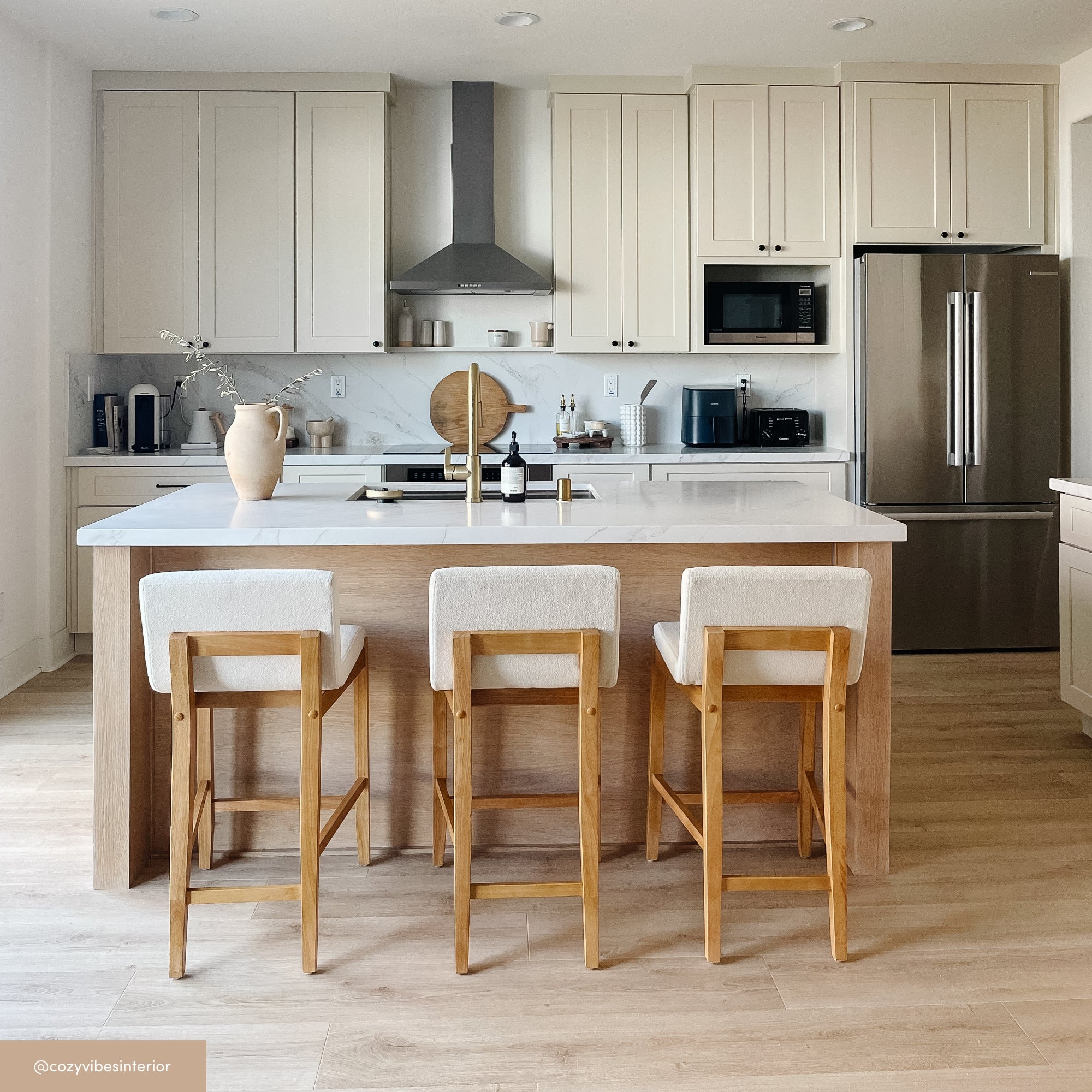 A modern kitchen with light wood floors, white cabinets, stainless steel appliances, and a large island with three light wood bar stools. Minimalist decor includes a vase, small plants, and neutral-colored accessories.