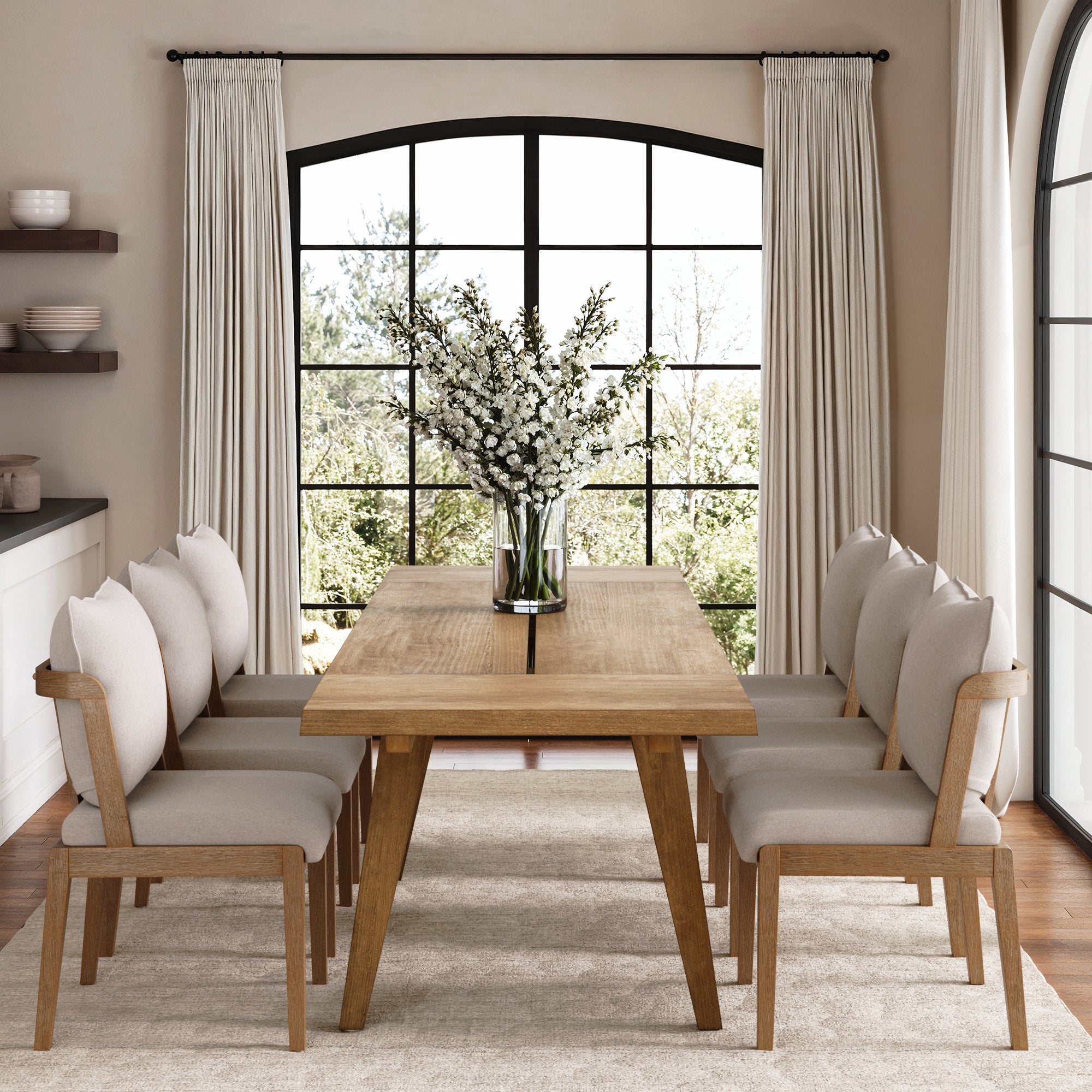 A bright dining room features a wooden table, a set of six Nathan James Scandinavian Dining Chairs with Back in Light Brown, a vase of white flowers, large windows with cream curtains, and a neutral rug on the floor.