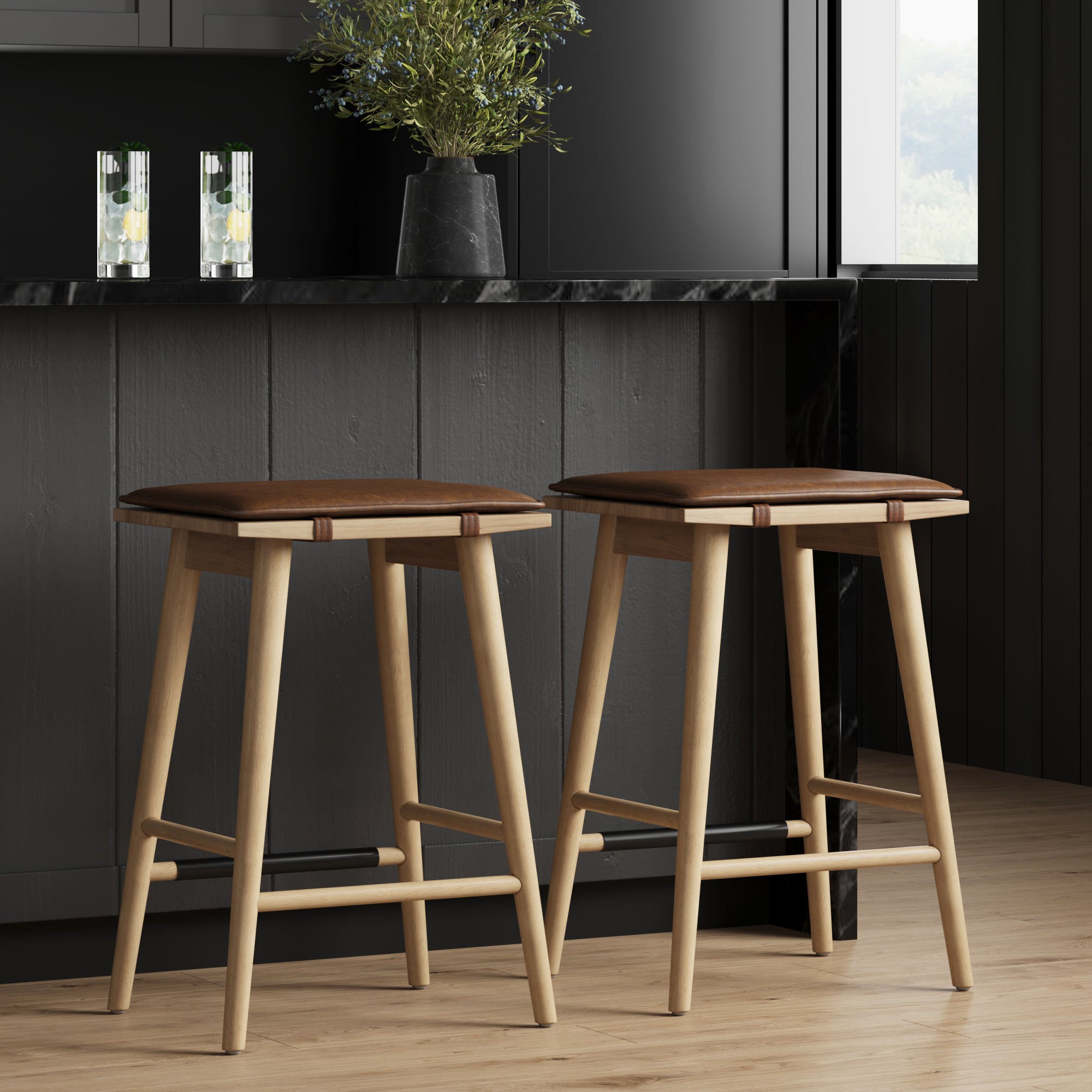Two wooden bar stools with brown seats are positioned at a modern dark kitchen counter. A vase with green foliage and two drinks are on the counter. The scene is well-lit with natural light from a window.