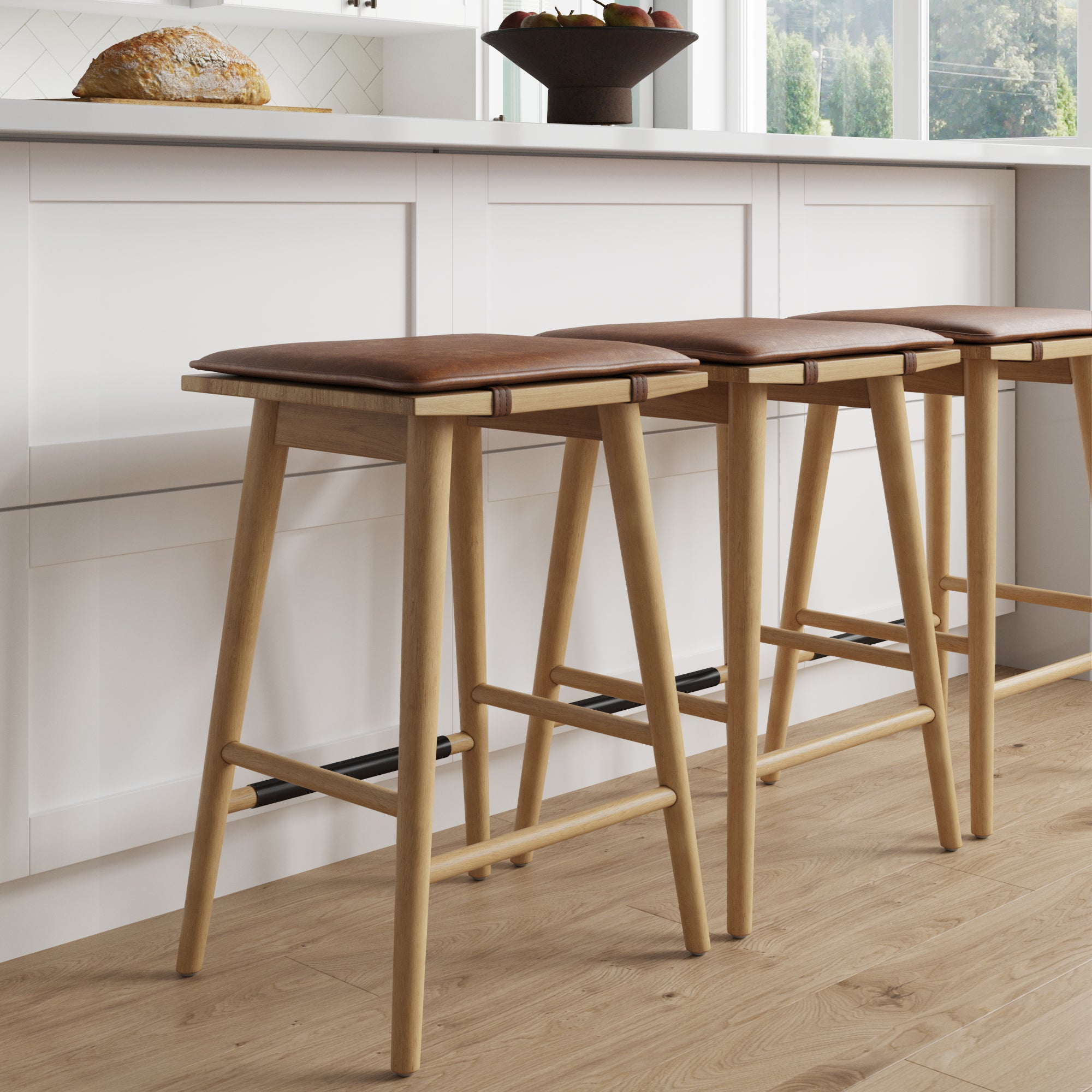 Three wooden bar stools with brown cushioned seats are lined up at a white kitchen island with a loaf of bread and a fruit bowl on the counter. Natural light streams in through large windows in the background.