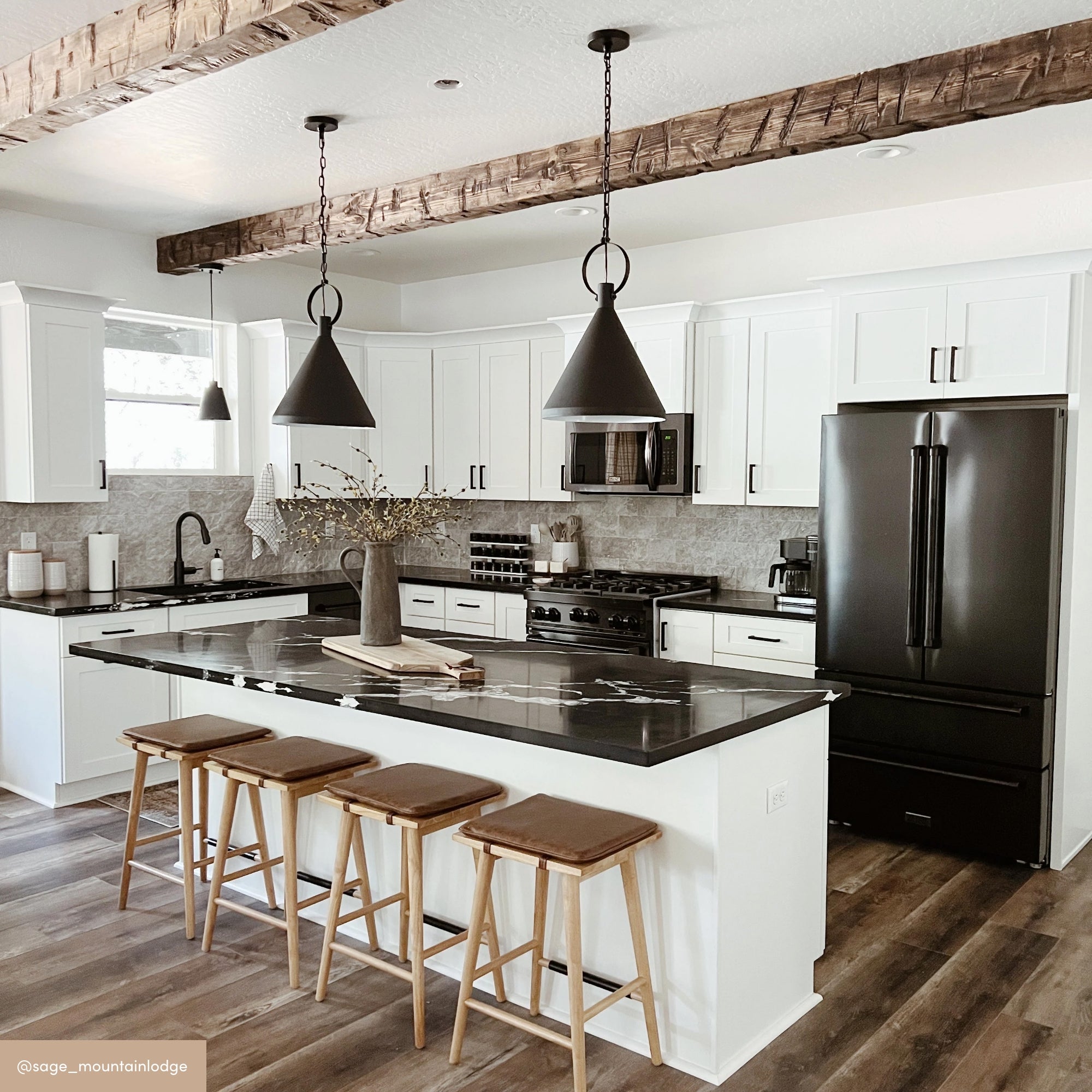 Modern kitchen with white cabinets, black appliances, a black marble island with wooden stools, pendant lights, wooden ceiling beams, and wood-style flooring. A vase with branches sits on the island.