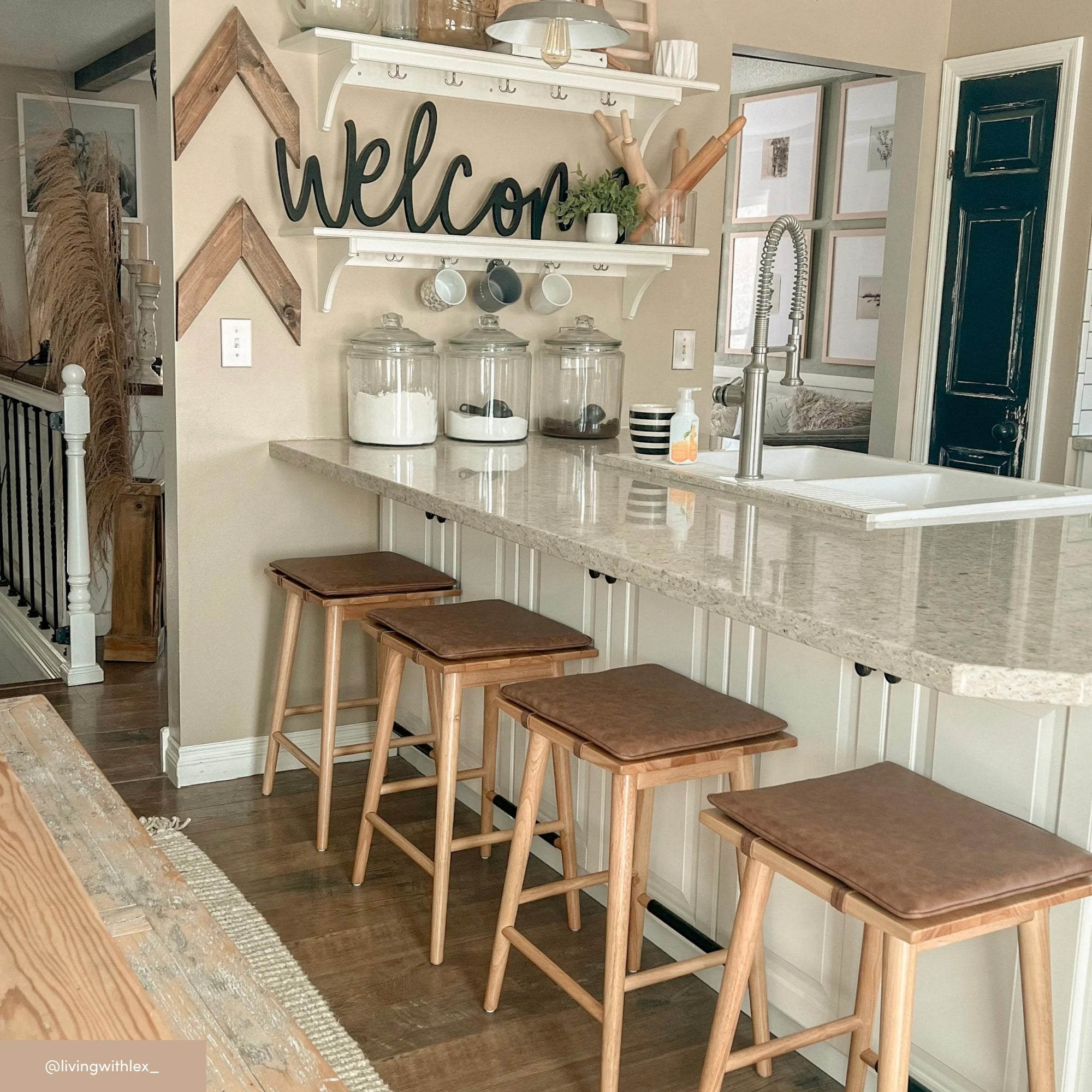 A bright kitchen with a marble countertop, four wooden stools, glass jars with ingredients, a sink, and a shelf with decor above. A large welcome sign and zigzag wall art add a cozy touch.