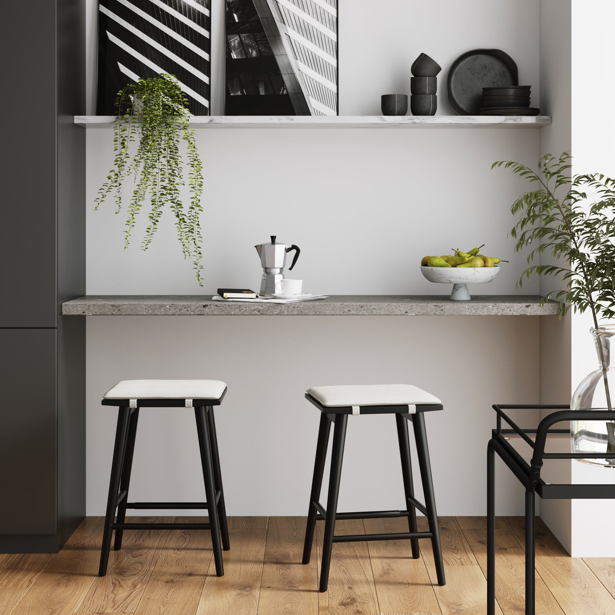 Minimalist kitchen nook with two Nathan James Boucle Counter Height Bar Stools in black, padded saddle seats at a concrete countertop, coffee maker, bowl of apples, potted plants, and monochrome art on a shelf against a white wall and wooden floor.