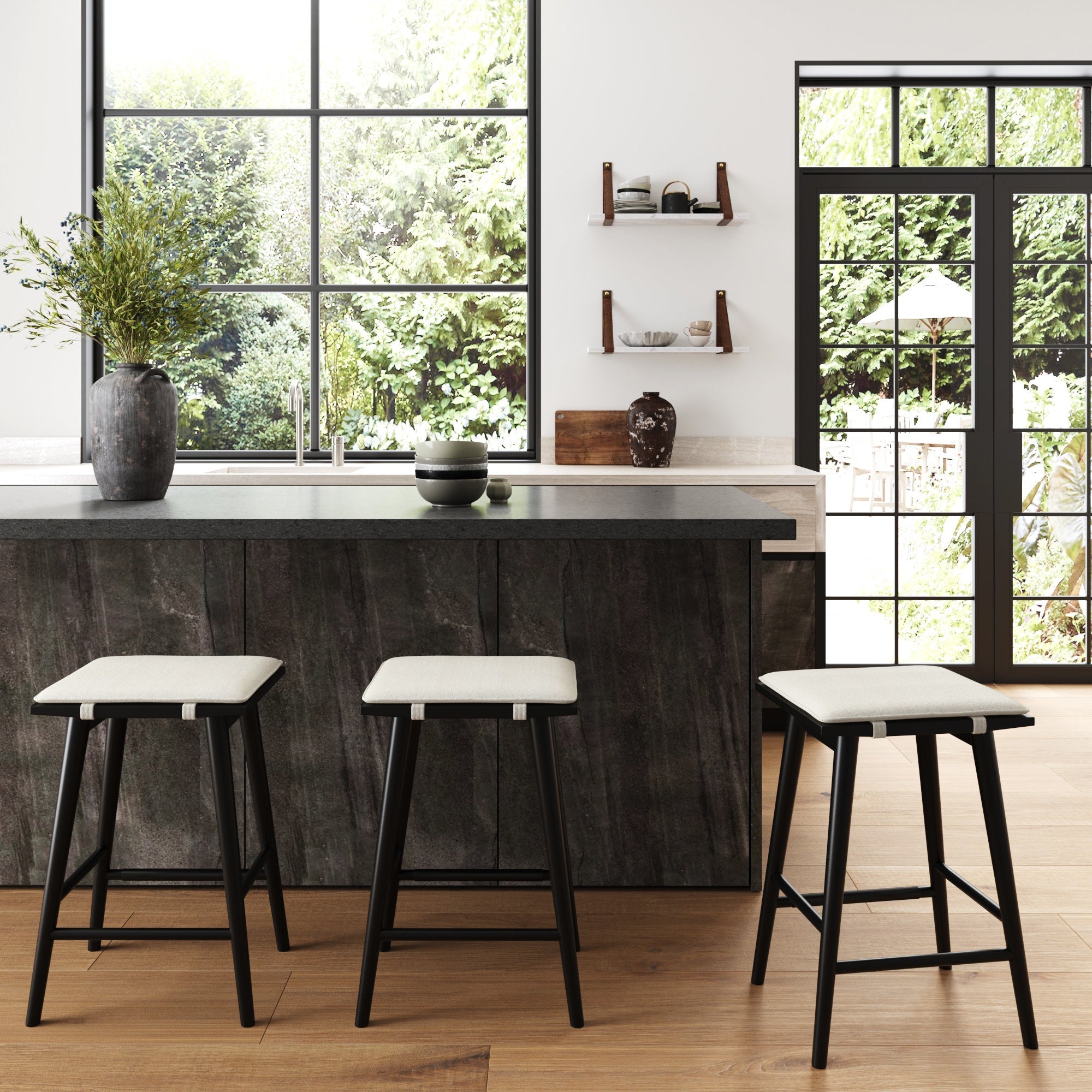 Modern kitchen with large windows, a dark island featuring three Nathan James Wood & Boucle Counter Height Bar Stools in black, potted plant on the counter, wooden floor, wall shelves with decor, and glass doors opening to a garden.