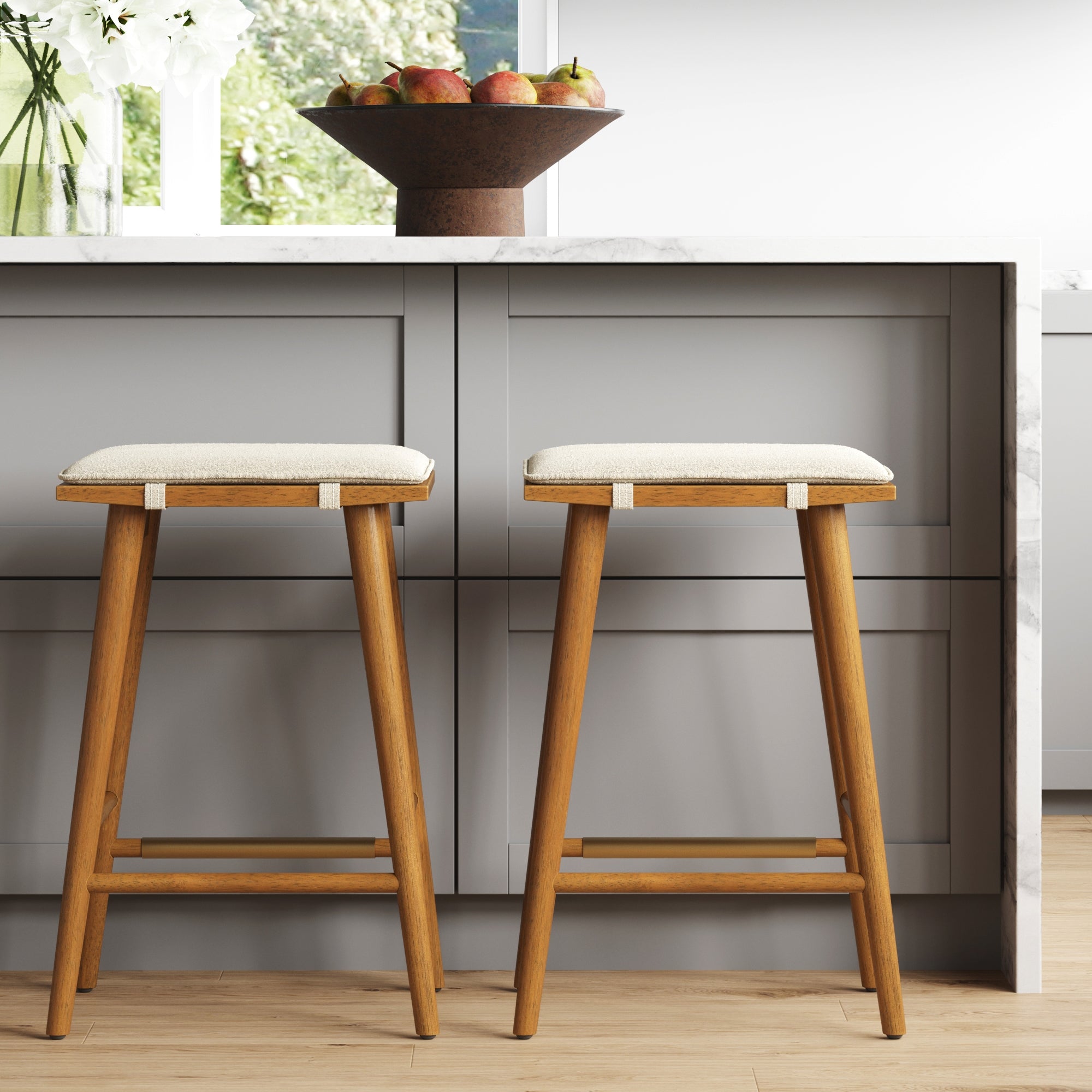 The Nathan James Boucle Counter Height Bar Stools in brown (set of 2) with padded saddle seats and solid wood frames are placed by a gray kitchen island topped with a bowl of apples, sunlight streaming through the window behind.