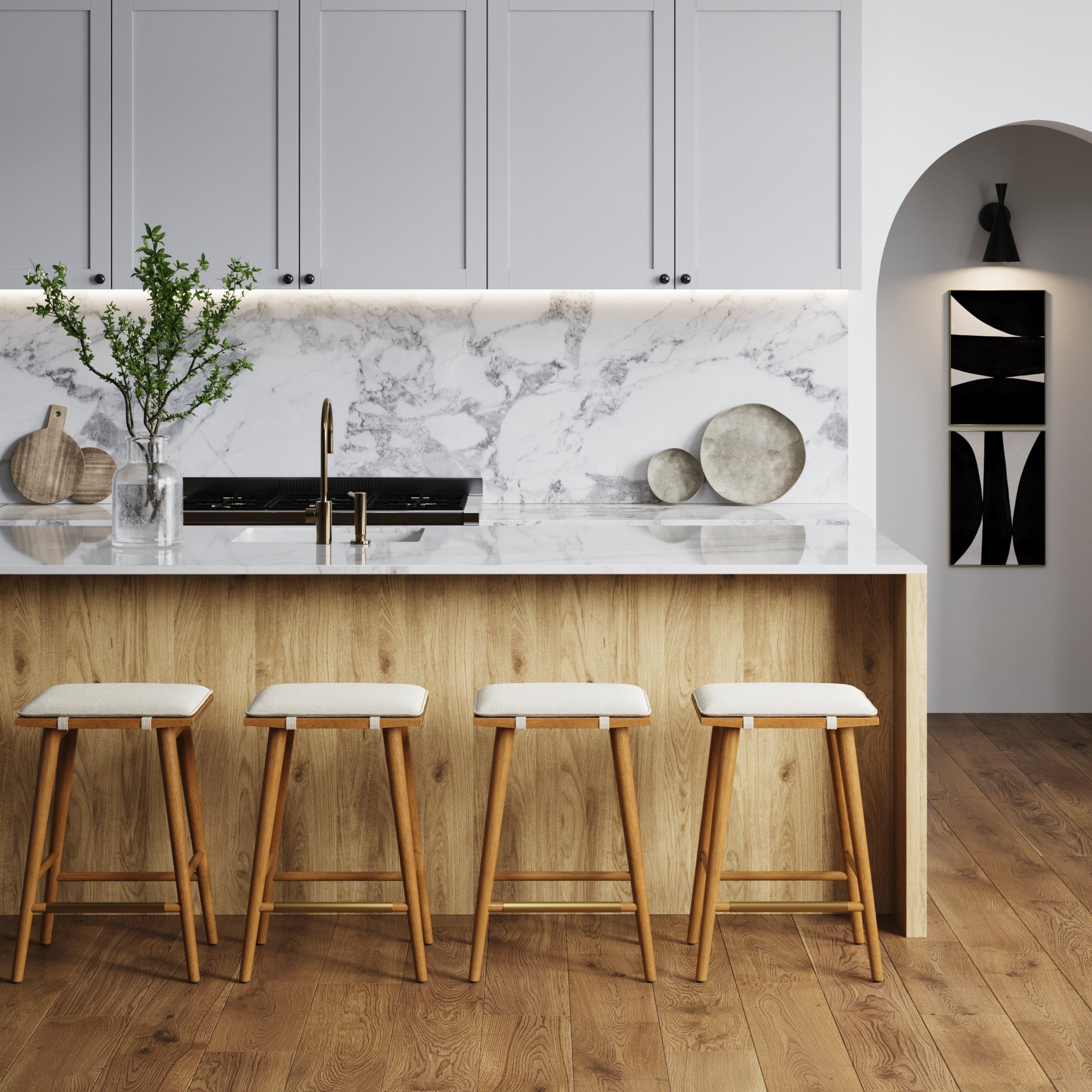 Modern kitchen with a marble backsplash, light wood island, and four Nathan James Boucle Counter Height Bar Stools in brown, featuring padded saddle seats, white upper cabinets, gold faucet, minimalist decor, a vase with branches, and abstract art.
