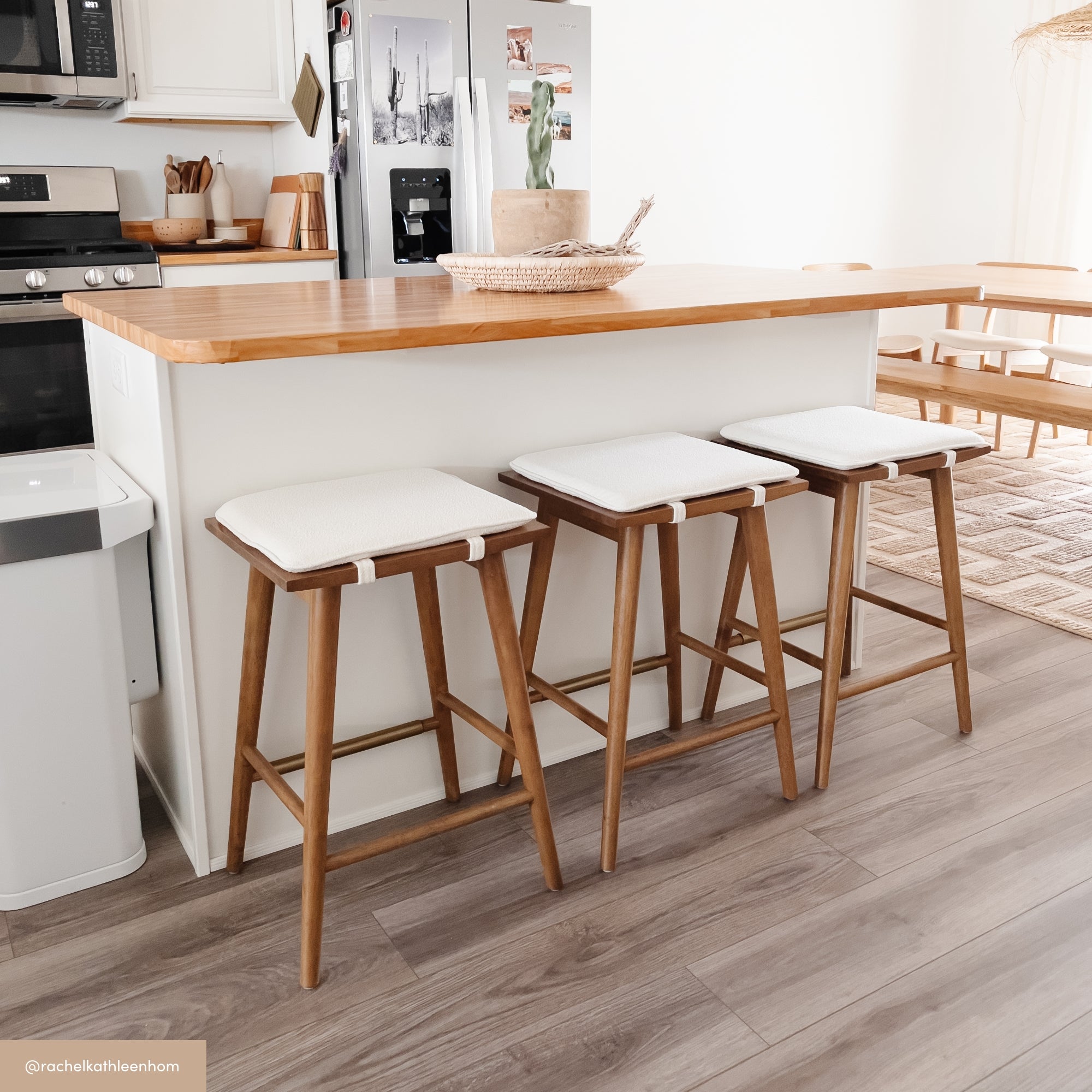 A modern kitchen with a wooden island countertop, three wooden barstools with white cushions, white cabinets, and light wood flooring. A dining table and rug are visible in the background.