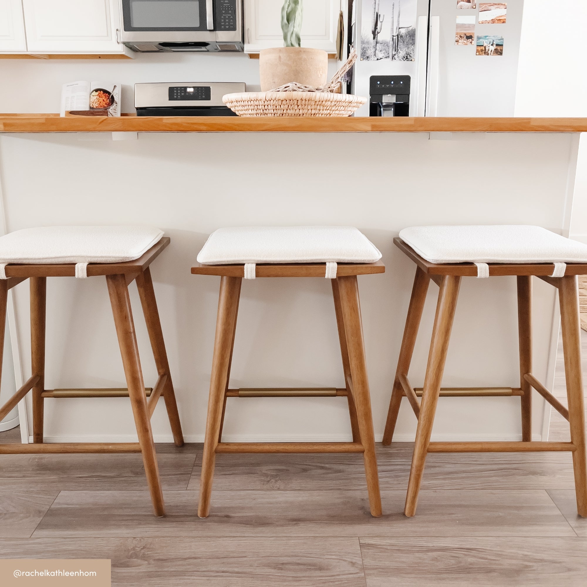 Three wooden bar stools with white cushioned seats are lined up in front of a white kitchen counter with a wooden trim. The background includes kitchen appliances and decor. Light wood flooring is visible.