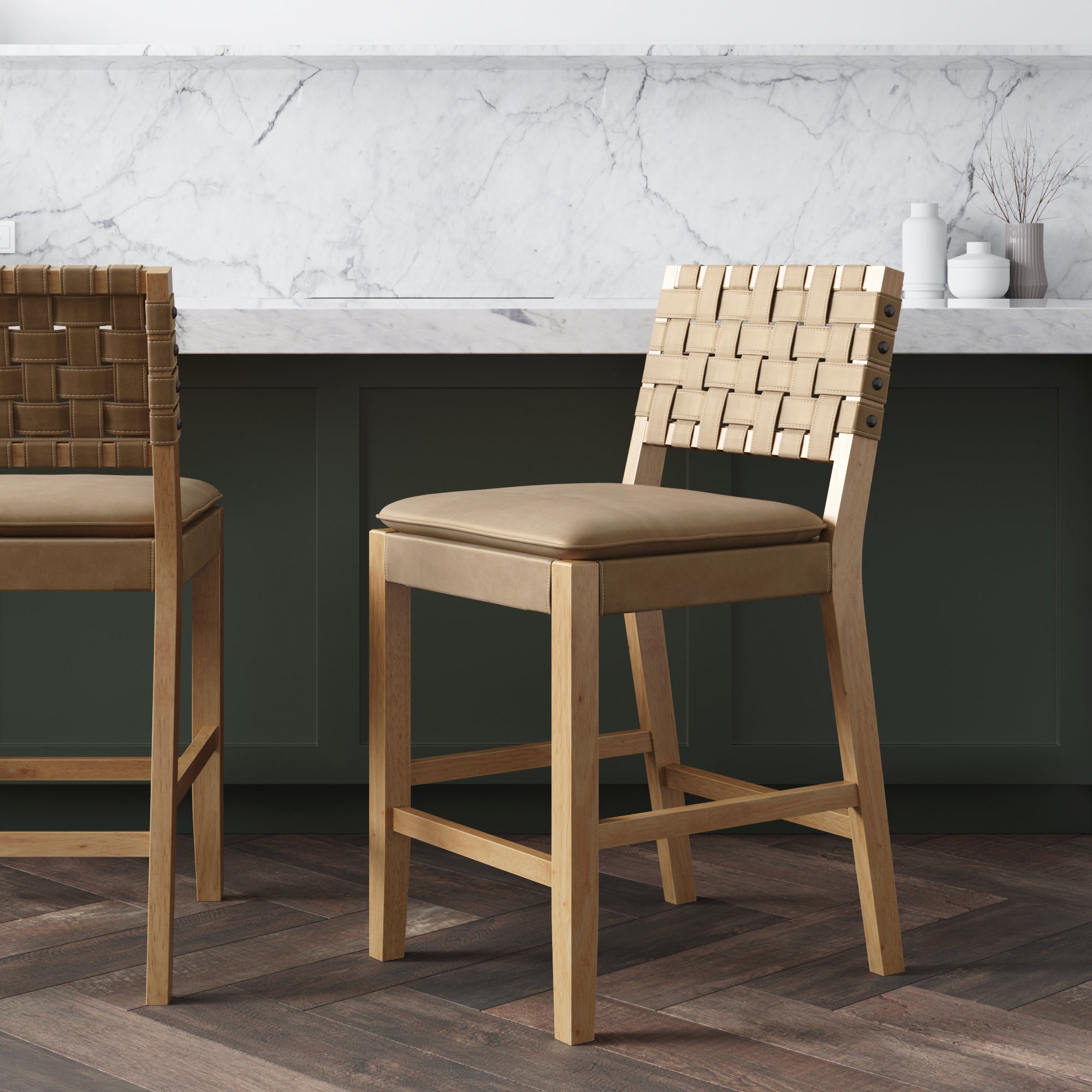 Two wooden counter stools with tan woven-backrests and padded seats sit in a modern kitchen with a marble backsplash, dark green cabinets, and dark herringbone wood floors.