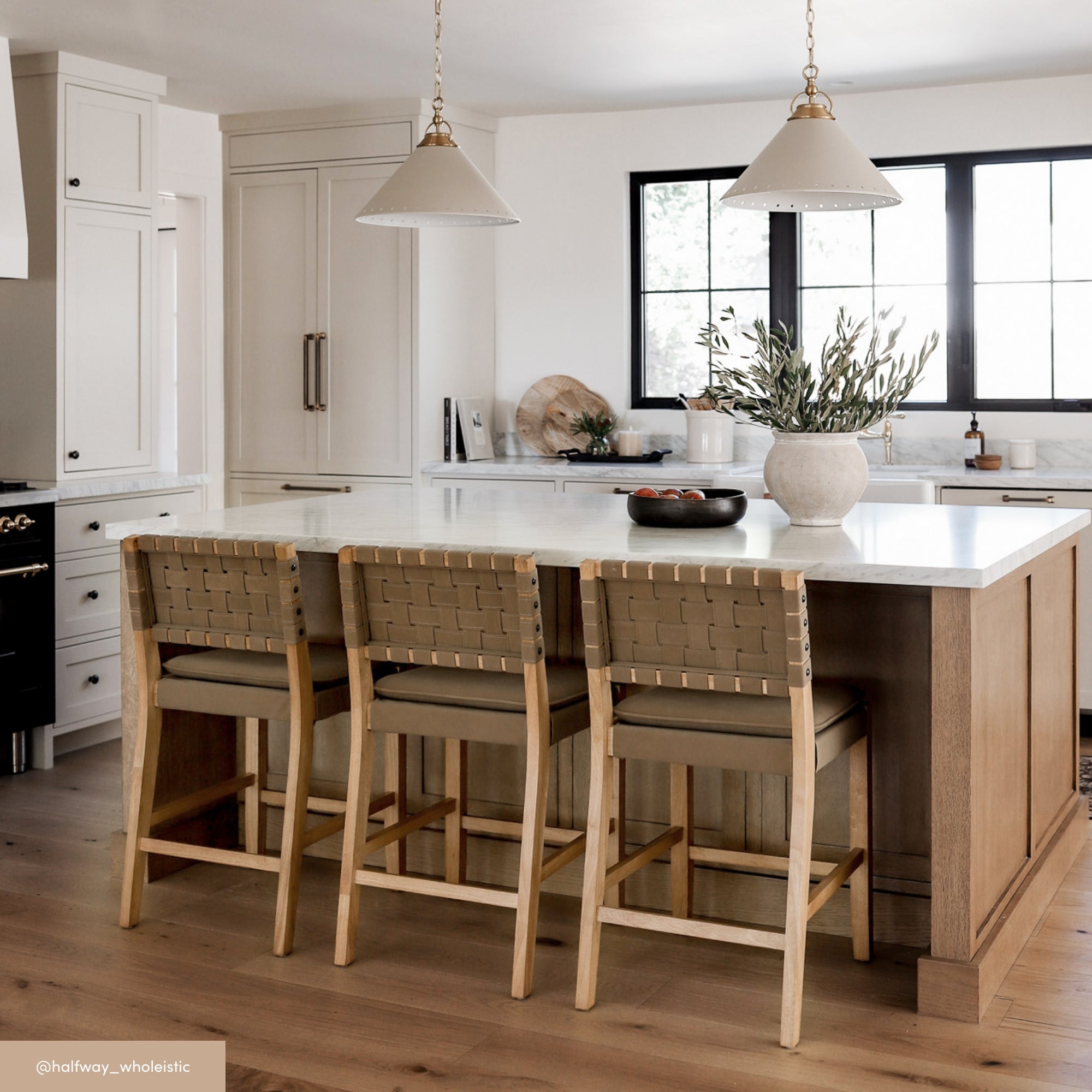 A modern kitchen with a large island featuring a white countertop, three wooden barstools with woven backs, pendant lights above, neutral cabinetry, and a vase with greenery on the counter.