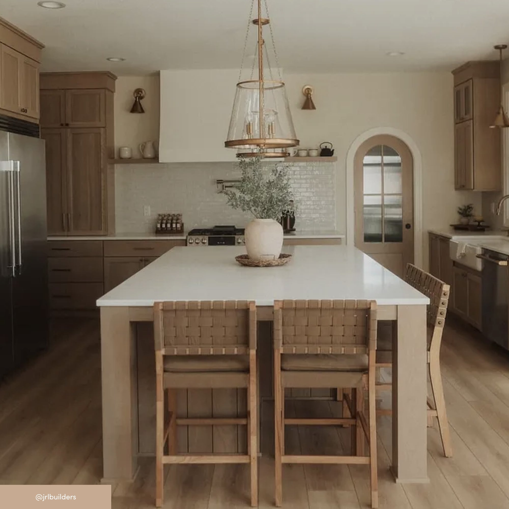 A modern kitchen with light wood cabinets, a large white island, woven bar stools, stainless steel appliances, and a decorative vase with greenery as a centerpiece; pendant lights hang above the island.
