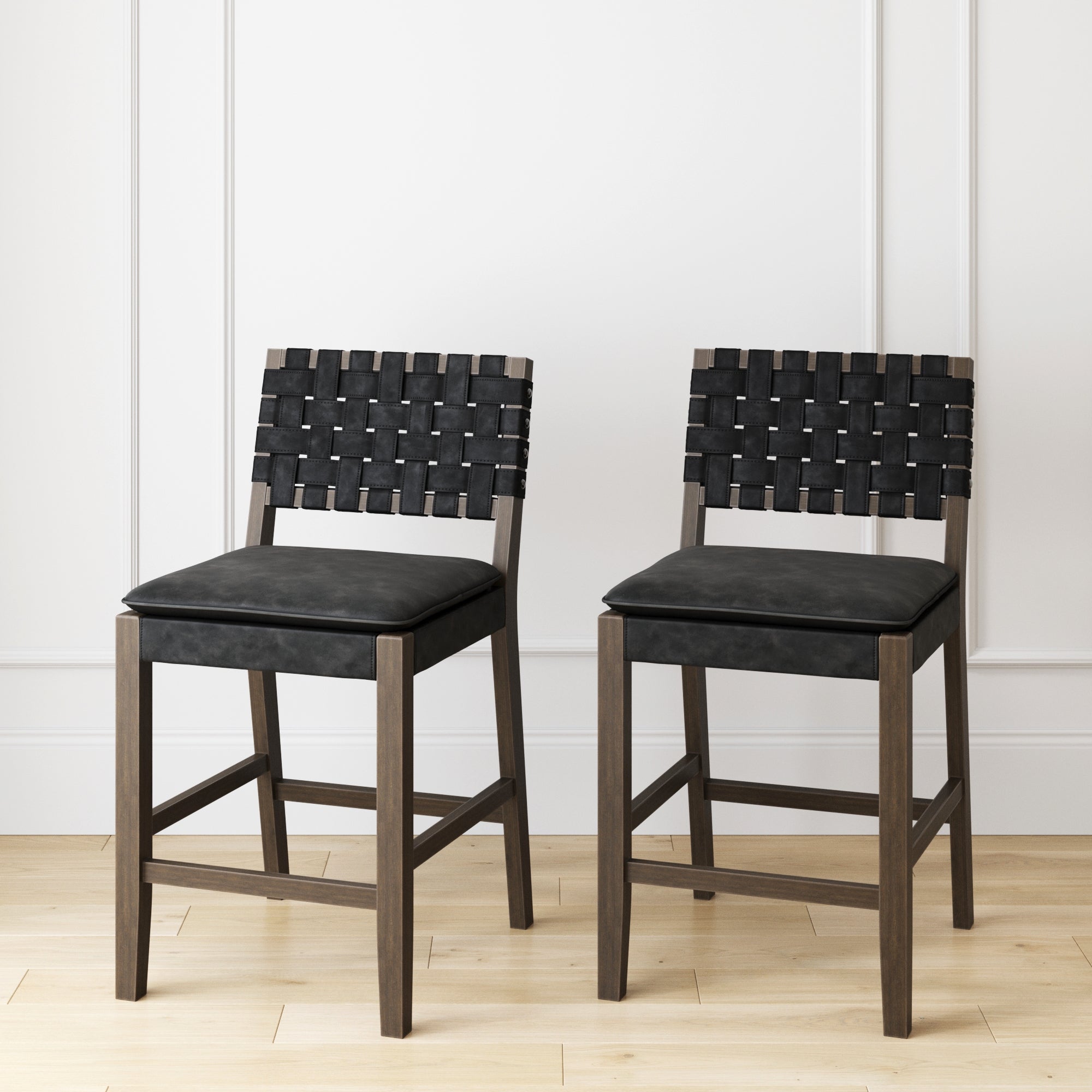 A set of two Nathan James Faux Leather Woven Counter Height Bar Stools in black, featuring solid wood legs and padded seats, stands on a light wood floor against a white paneled wall.