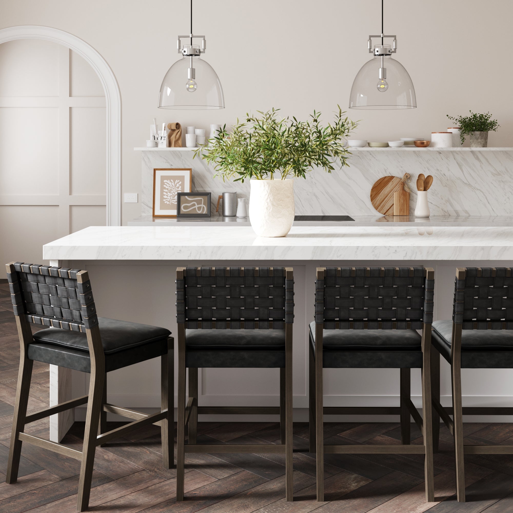 Modern kitchen with a marble island, three black woven bar stools, a large white vase with green foliage, pendant lights above, and minimalist decor on white shelves in the background.