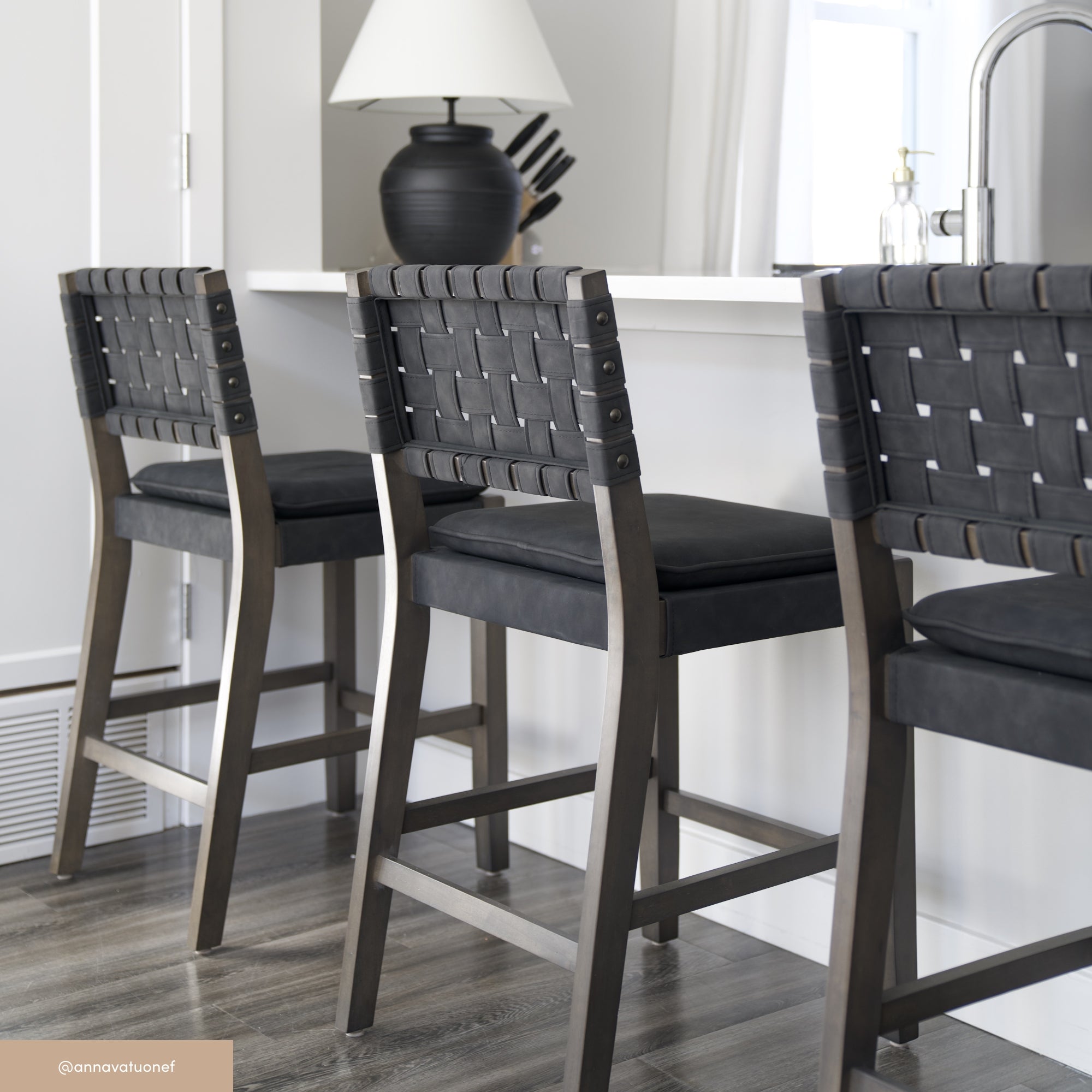 Three modern barstools with woven black leather backs and cushioned seats are lined up at a white kitchen counter. A black lamp, a white vase, and a window are visible in the background.
