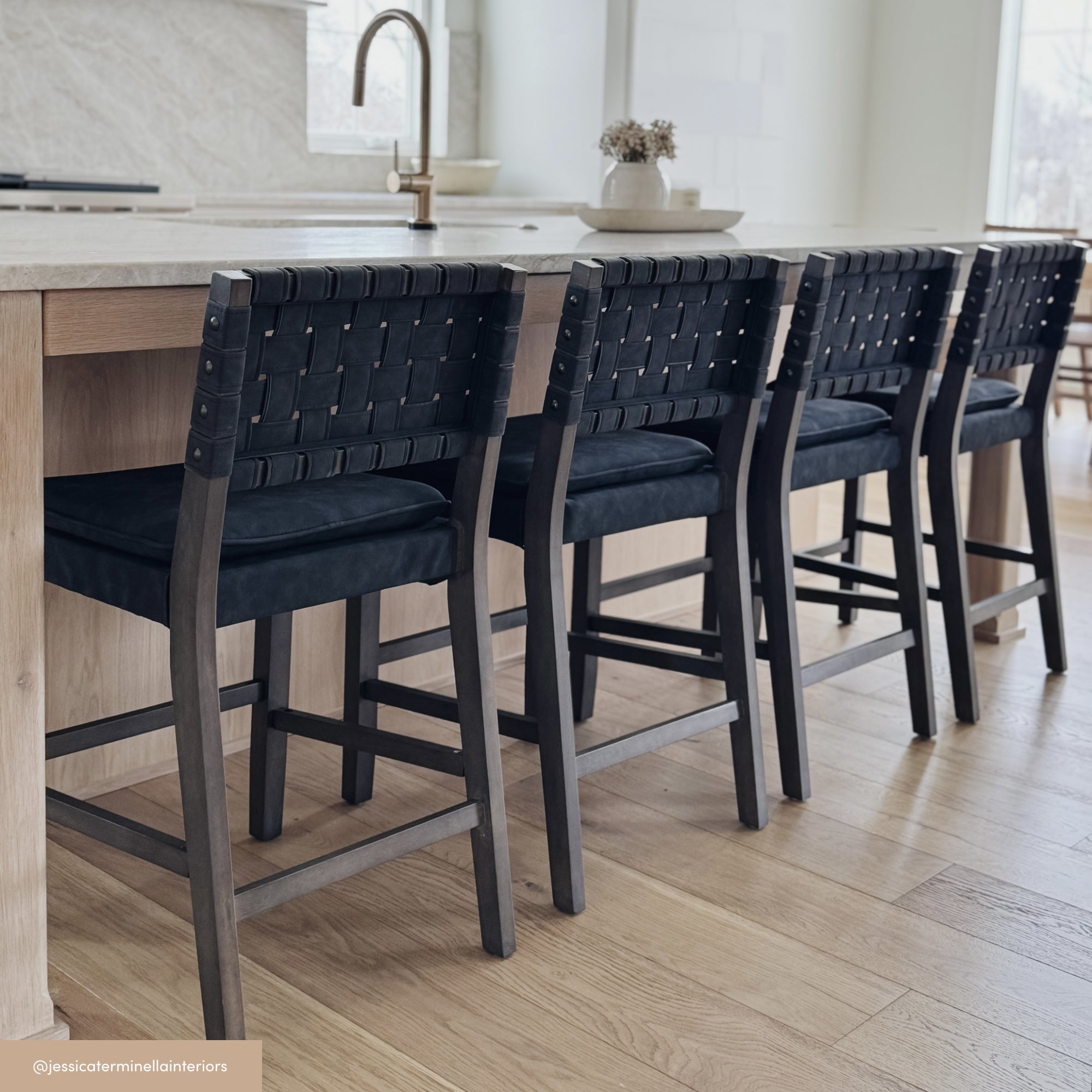 Four black woven-back counter stools are lined up along a light wood kitchen island with a white countertop. The kitchen features light wood flooring and neutral, minimalist decor.