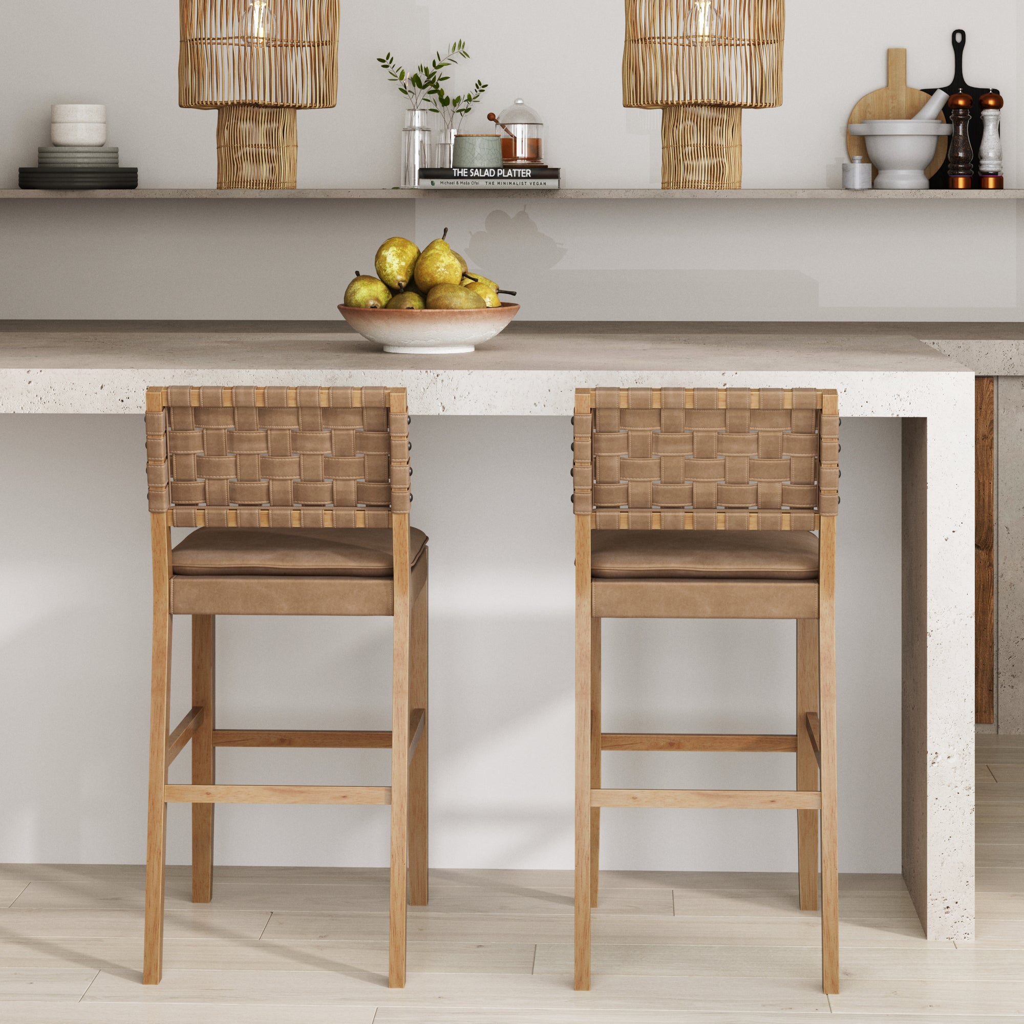 Two woven tan barstools sit at a light stone kitchen island. On the counter is a bowl of pears. The background features shelves with decorative items, plants, and woven pendant lights.