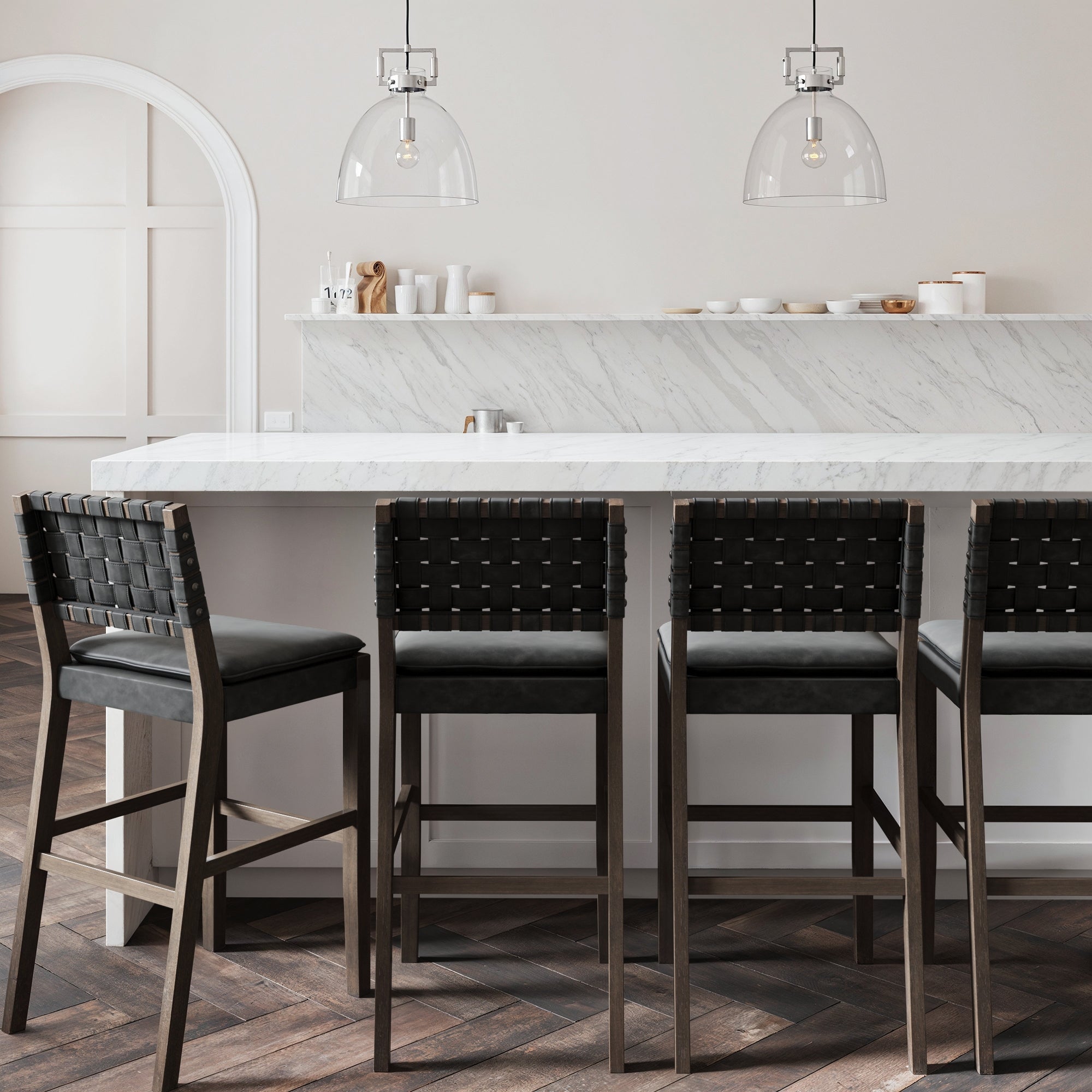 A modern kitchen features a white marble island, four Nathan James Faux Leather Woven Bar Stools in black, pendant lights, and minimalist decor on open shelves set against a white and marble backsplash.