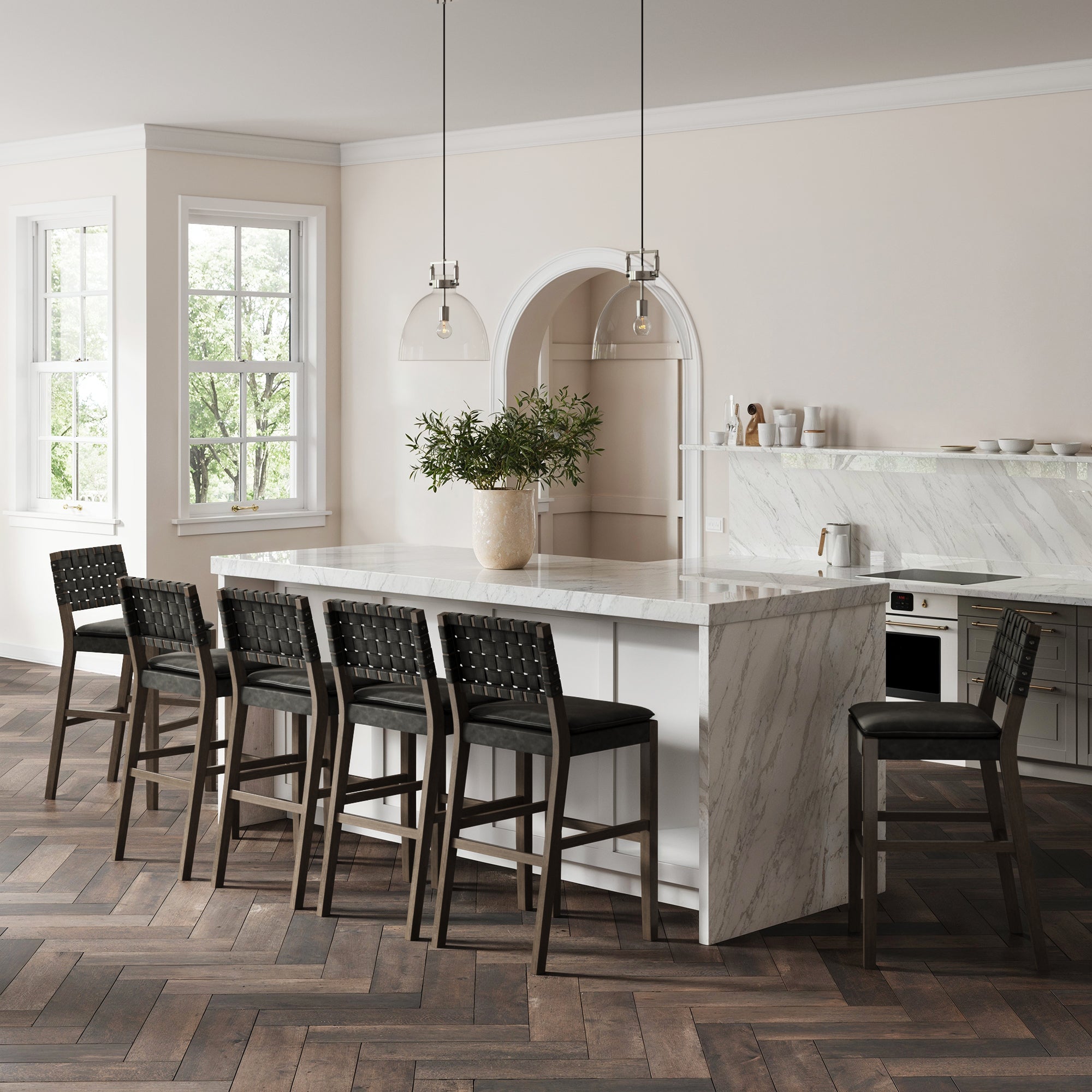 Modern kitchen featuring a large marble island, six Nathan James Faux Leather Woven Bar Stools in black, wooden herringbone floor, white walls, large windows, a potted plant centerpiece, and pendant lights above the island.