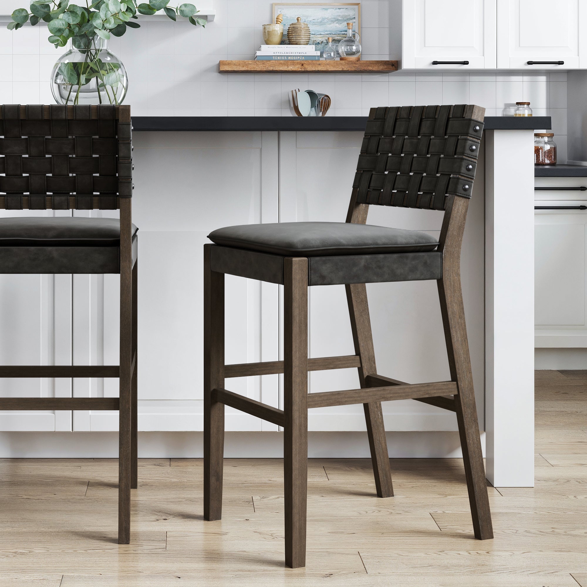 A modern kitchen showcases the Nathan James Faux Leather Woven Bar Stool Black, featuring a woven backrest and black cushioned seat at a white island, with another matching stool partially visible on the left.