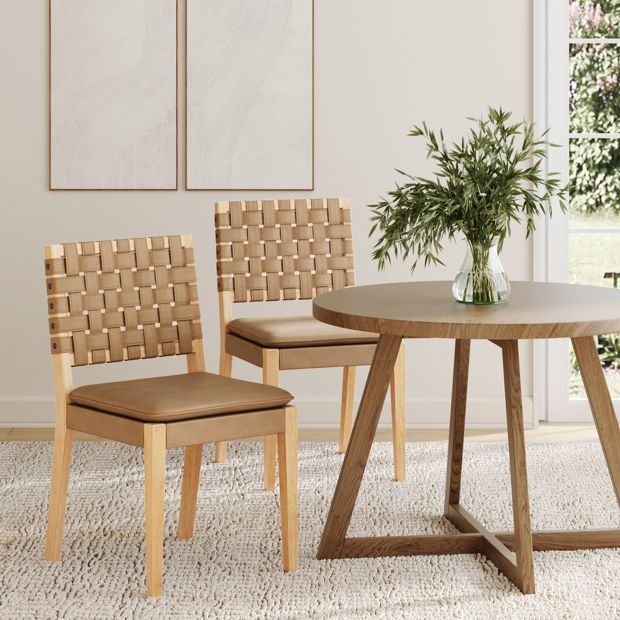 Two Nathan James Faux Leather Dining Chairs in light brown, featuring hand-woven backs and tan cushions, sit beside a round wooden table. Neutral walls, abstract art, a textured rug, and natural light complete the inviting room scene.
