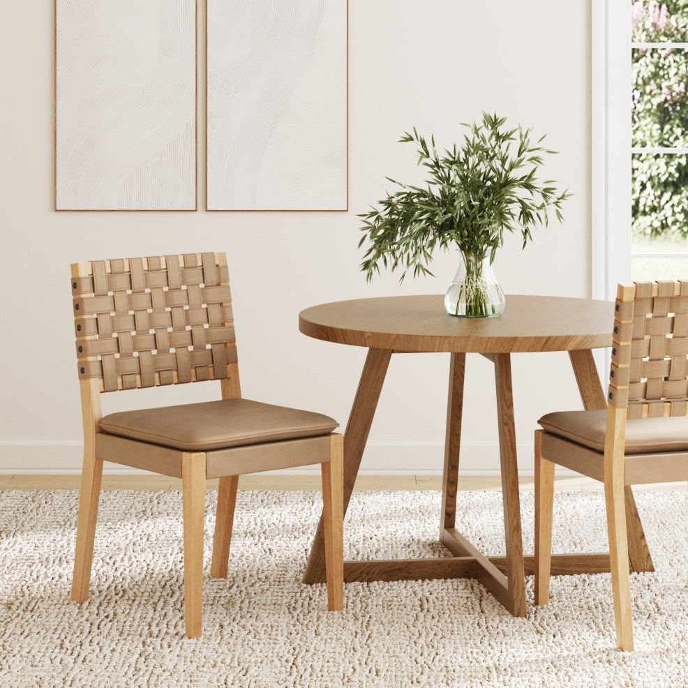 A round wooden table with a vase of greenery sits on a textured rug, flanked by two Nathan James Faux Leather Dining Chairs in light brown, in a bright, minimal room with abstract wall art and sunlight streaming through the window.