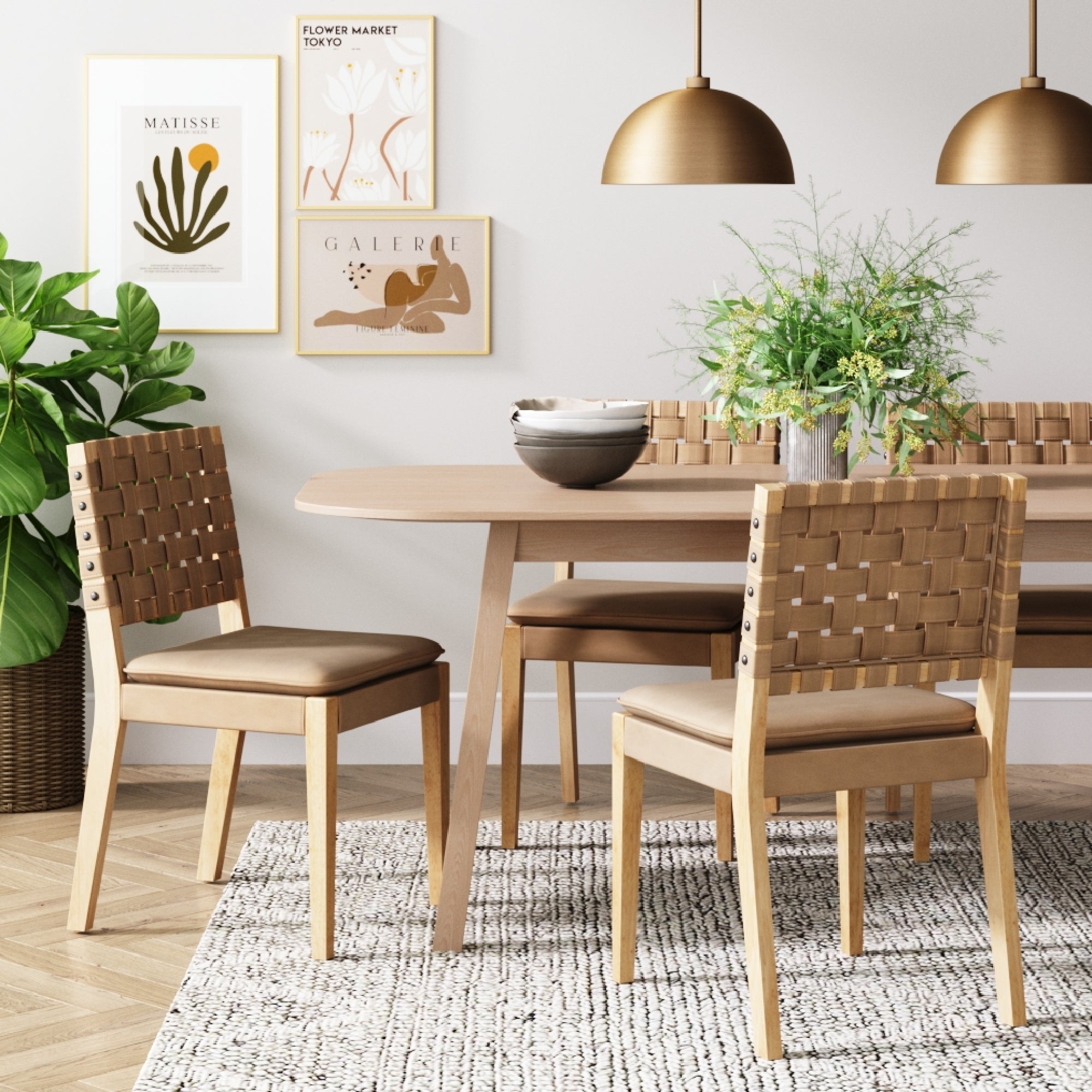 A modern dining room features a light wooden table, Nathan James Woven Faux Leather Dining Chairs in light brown with solid wood frames, a green plant, framed wall art, two gold pendant lights, and a textured rug on wooden floors.