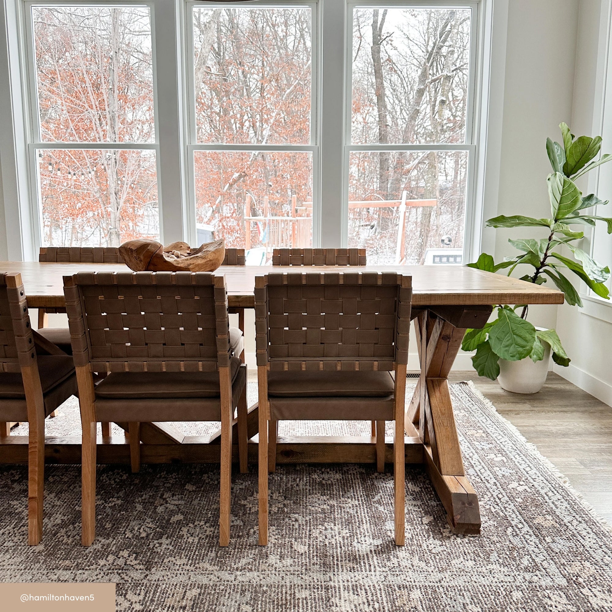 A wooden dining table with woven chairs sits on a patterned rug in front of large windows. Outside, trees with orange leaves and snow are visible. A potted plant is in the corner of the bright, airy room.