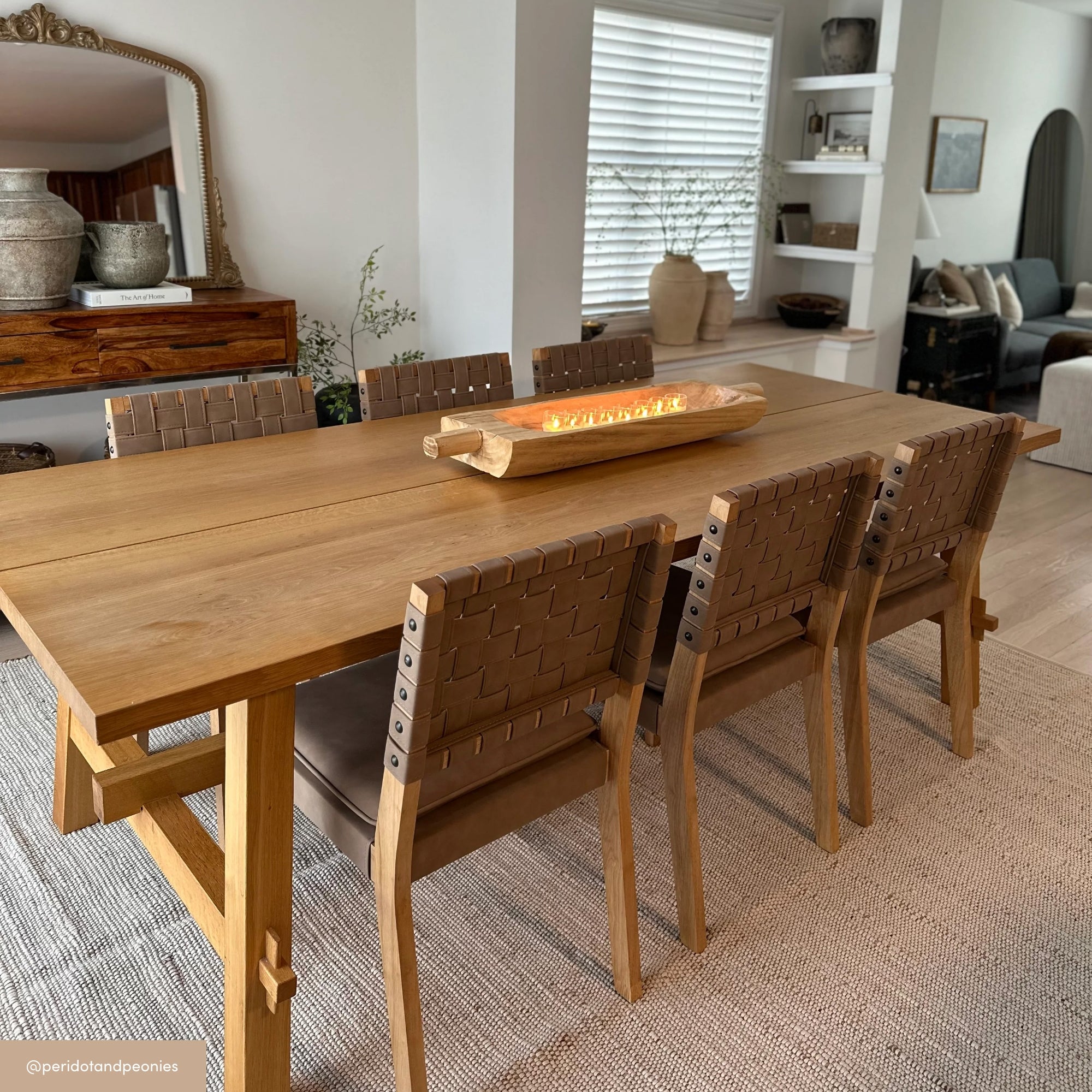 A modern dining room with a wooden table, six woven leather chairs, a rectangular wooden centerpiece, neutral tones, a textured rug, and decor items on shelves and sideboard in the background.