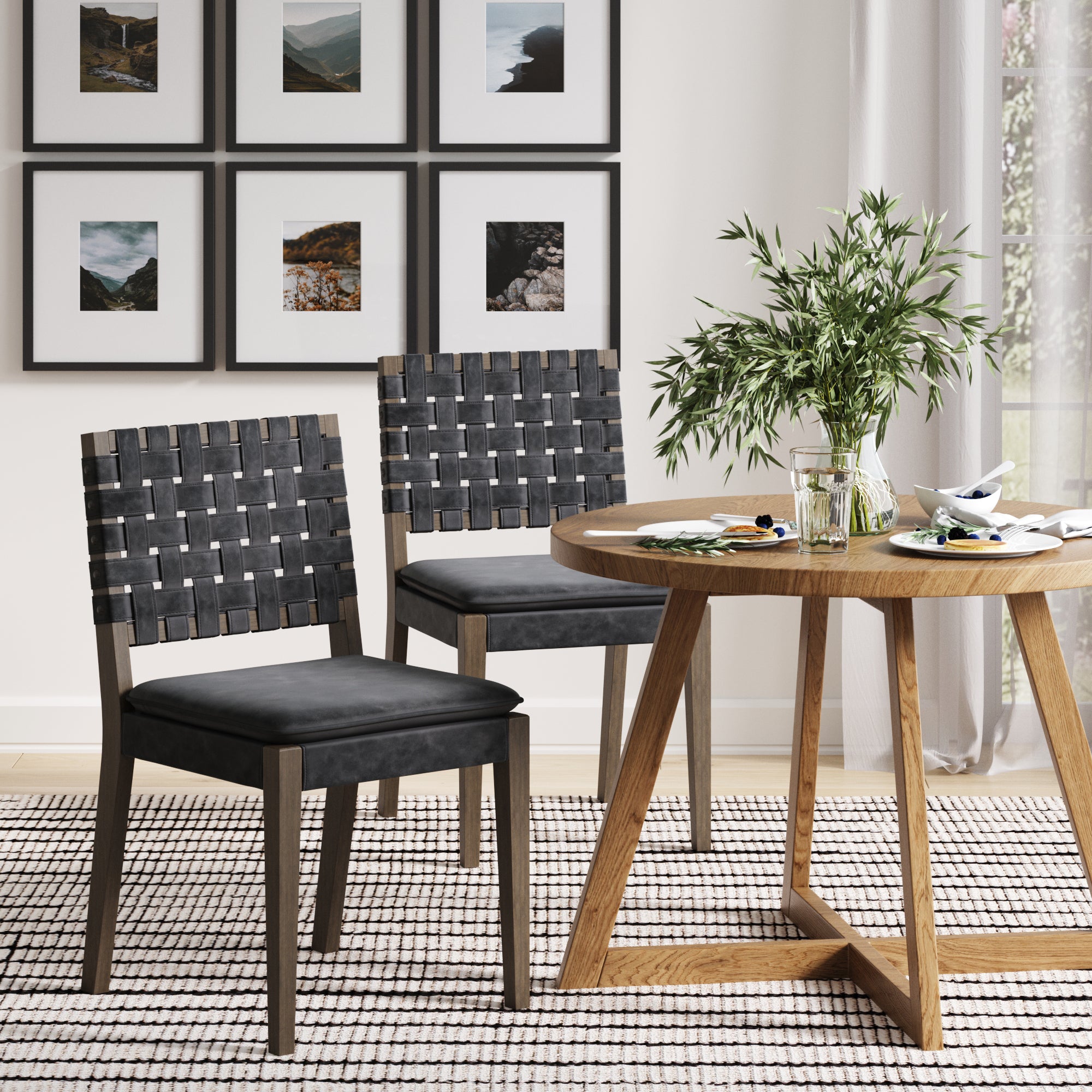 A modern dining area with a round wooden table, a plant centerpiece, and a gallery wall. Two Nathan James Faux Leather Dining Chairs (Black, Set of 2) with solid wood frames sit on a striped rug under natural light from the window.