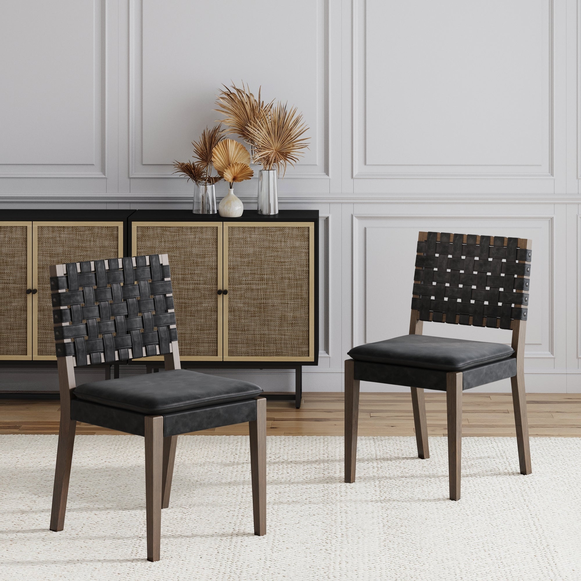 Two Nathan James Faux Leather Dining Chairs in black rest on a light rug. Behind them, a sideboard with cane doors and decorative vases with dried plants sits against a white paneled wall.