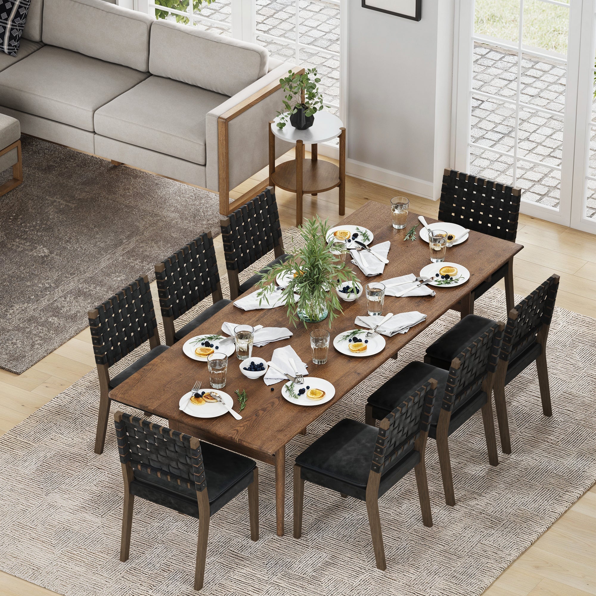 A Nathan James wooden dining table is set for a meal with plates, glasses, napkins, a green centerpiece, and eight black faux leather dining chairs (Set of 8) featuring solid wood frames. The table sits on a rug in a bright room with large windows and a sofa.